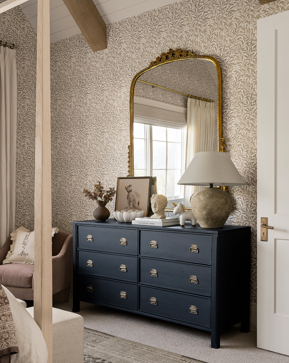 A cozy bedroom corner features the Mackenna Dresser with elegant brass pulls, topped with ceramic decor, a table lamp, and framed art. A large ornate gold mirror, light wallpaper, and soft furnishings complete the inviting atmosphere.