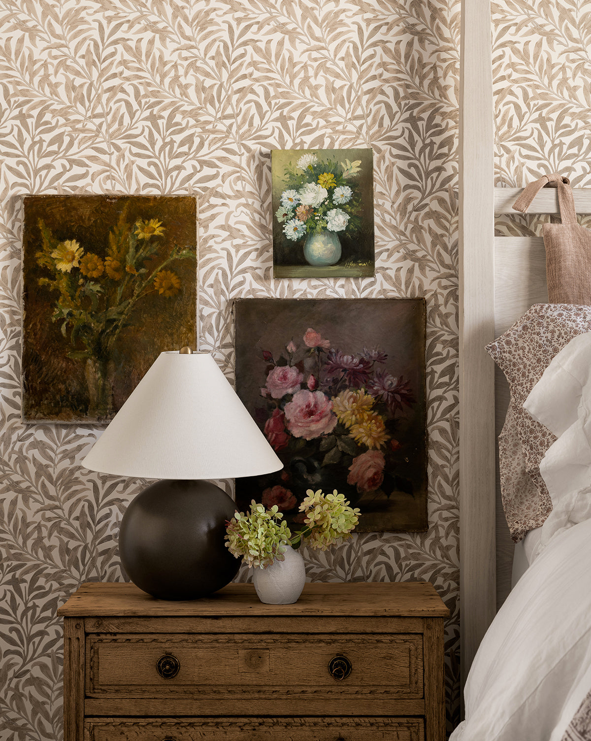 A cozy bedroom corner features the Tess Ceramic Table Lamp on a wooden nightstand, with a small flower vase and three floral paintings set against leafy wallpaper beside a white bed, by McGee & Co.