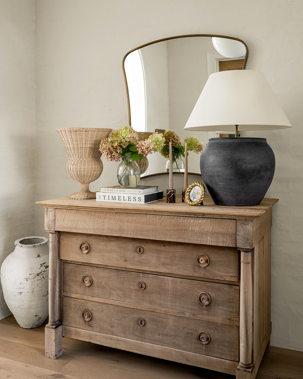 A rustic wooden dresser with three drawers displays a black lamp, wicker basket, floral vase, books, gold clock, and the Ludwig Wall Mirror. Beside the dresser on the floor is a large decorative ceramic pot, by McGee & Co.