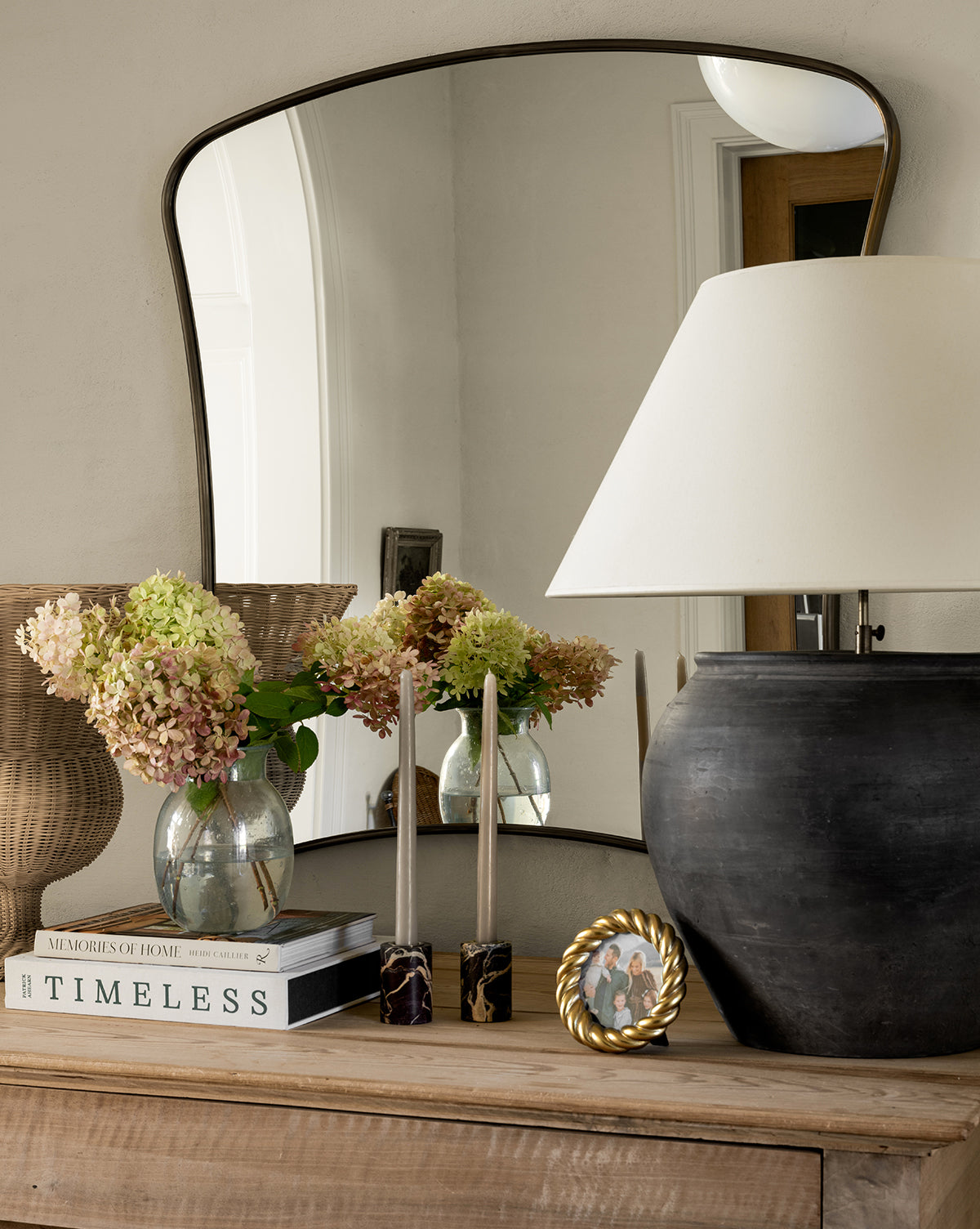 A wooden console table features stacked books, a vase of hydrangeas, Perrin Marble Taper Holders (Set of 2), a round picture frame, a large black lamp with white shade, and a curvy mirror reflecting part of the room.
