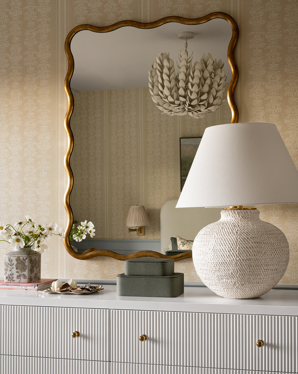 A dresser with gold knobs displays the Avedon Table Lamp with a white linen shade, two green boxes, and a vase of white flowers. Above, a large wavy gold-framed mirror reflects light fixtures and part of the room, by McGee & Co.