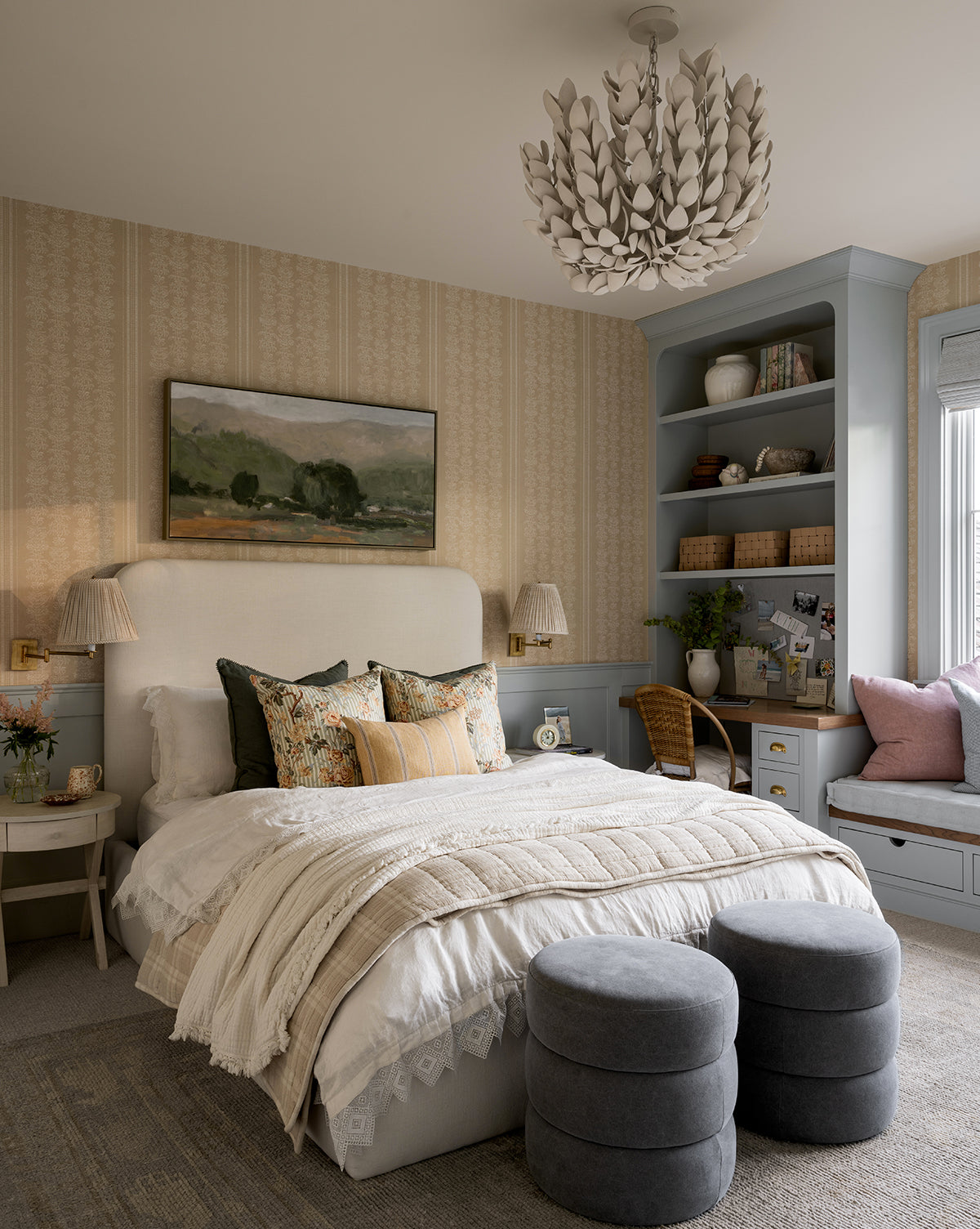 A cozy bedroom features a neutral upholstered bed with layered pillows and a quilted cream throw. Two Channel Ottomans in gray are at the foot of the bed, while built-in shelves, a desk, and a window seat with pink cushions complete the space by McGee & Co.