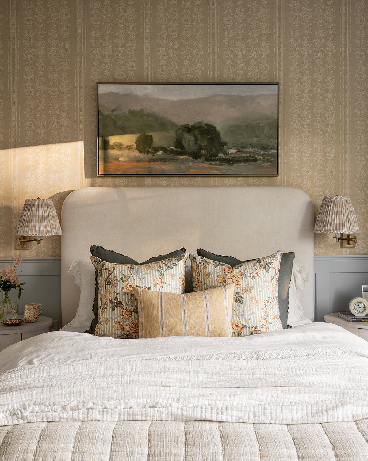 A cozy bedroom featuring the Southern June beige upholstered headboard, three decorative pillows, a white comforter, and two wall-mounted lamps. Abstract wall art with rolling hills in soft neutrals hangs above the bed.