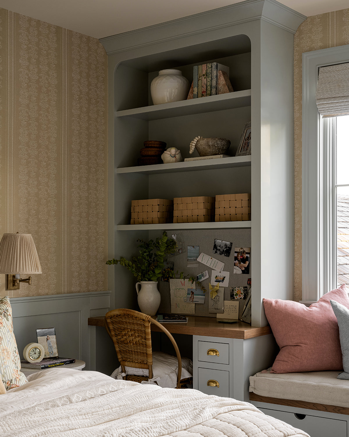 A cozy bedroom corner showcases a built-in desk with shelves, books, and decor. At the desk sits a woven leather chair and the Rupert Leather Bin for storage, all beside plants, pinned notes, a window seat with pillows, and a quilted bed.