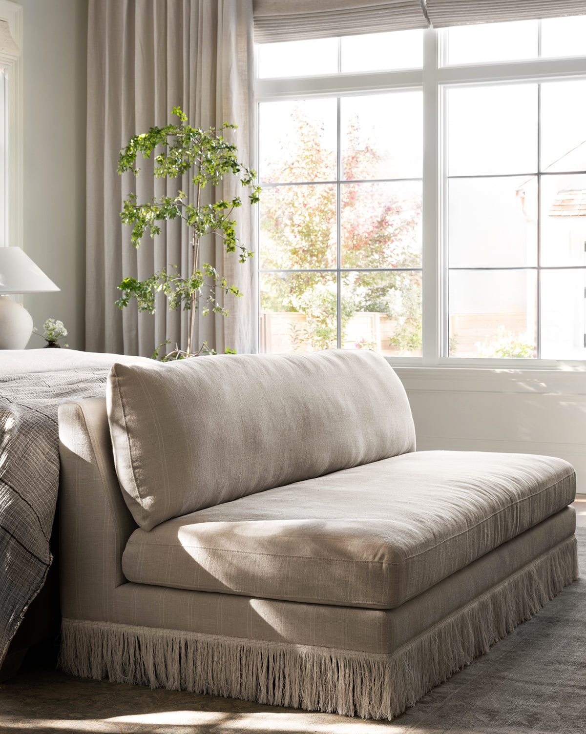 The Philippa Settee (Ready to Ship) with fringe trim sits by a sunlit window, next to a neatly made bed and tall potted plant, creating a cozy, serene bedroom corner, by McGee & Co.