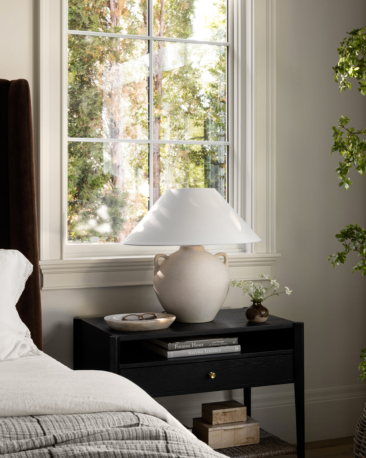 A cozy bedroom scene features a neatly made bed next to a black nightstand topped with the Mays Table Lamp, a dish, stacked books, and a small vase of flowers, all set before a sunlit window overlooking trees by McGee & Co.