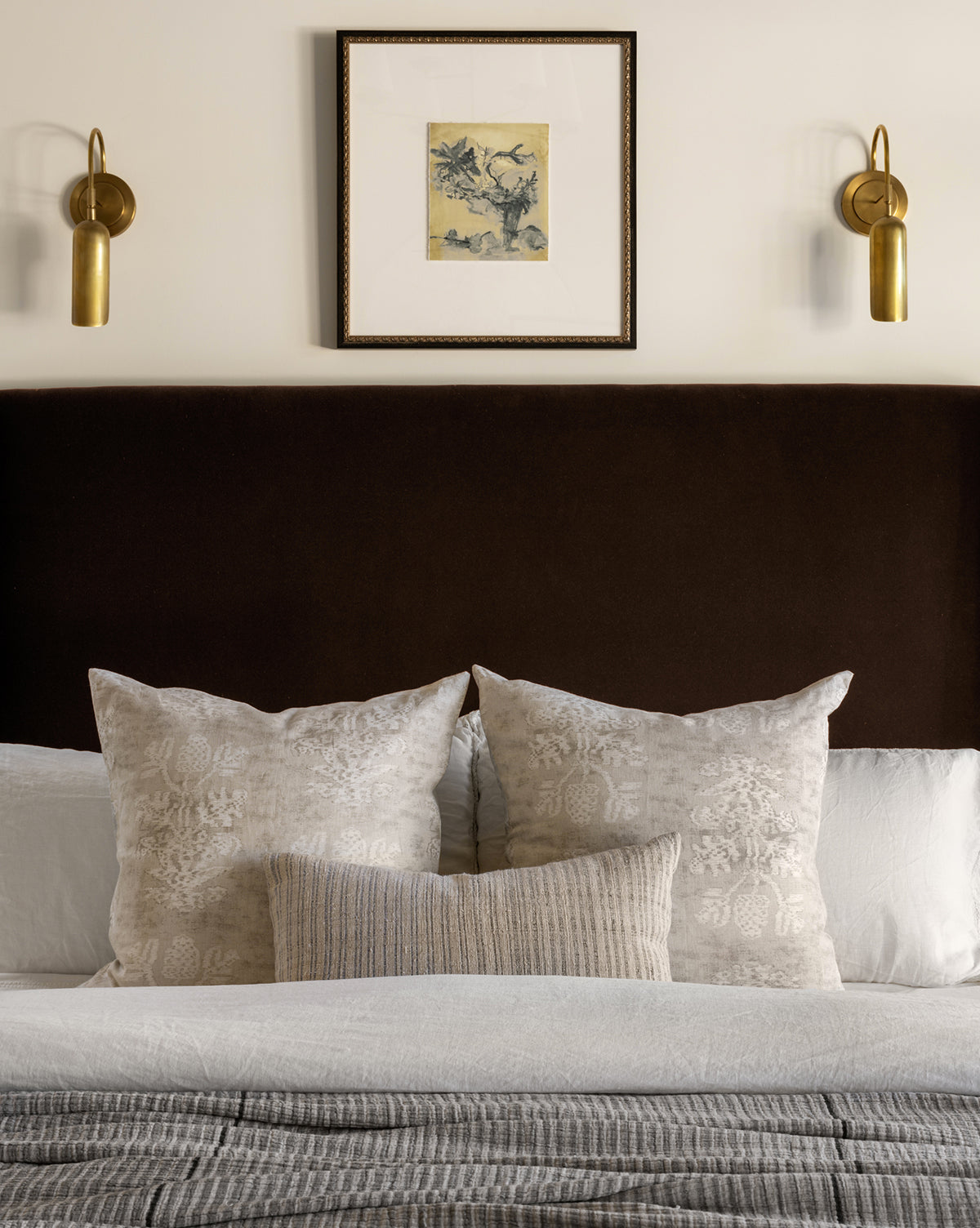 A neatly made bed with textured neutral pillows, a dark upholstered headboard, and two brass wall sconces sits beneath the Abstracted Votives botanical wall art, framed above the bed on a light-colored wall.