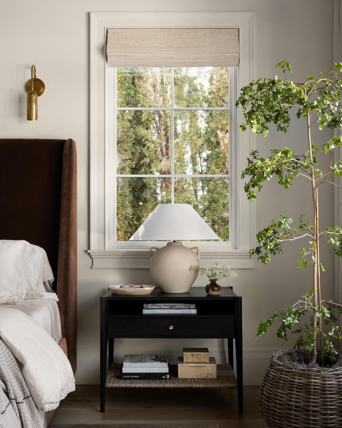 The Dana Nightstand, adorned with books, a ceramic vase, and a lamp, stands beside a bed with white bedding. Behind it is a window revealing green trees. To the right is a large potted plant; to the left hangs a brass wall sconce, by McGee & Co.