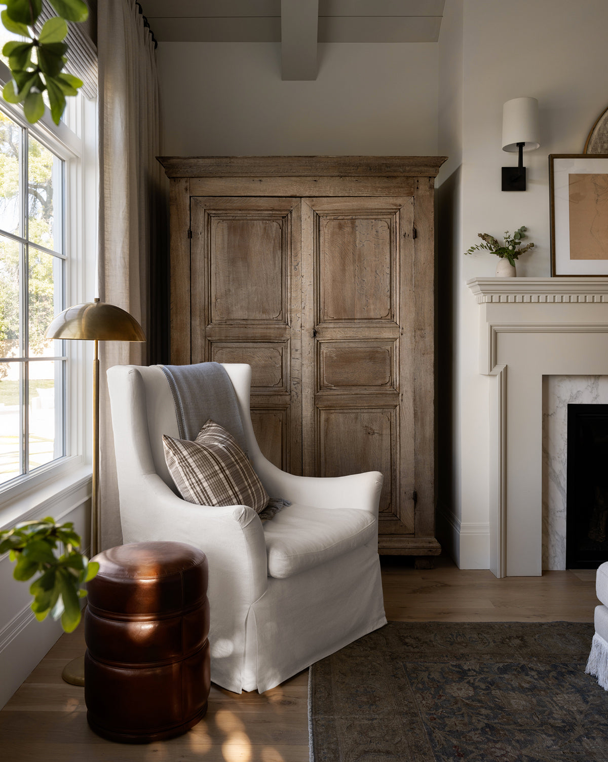 A cozy corner features a white armchair, plaid and striped pillows, a brown leather ottoman, the elegant Aldorno Floor Lamp, a large wooden armoire, window, and a white fireplace adorned with decor, by McGee & Co.