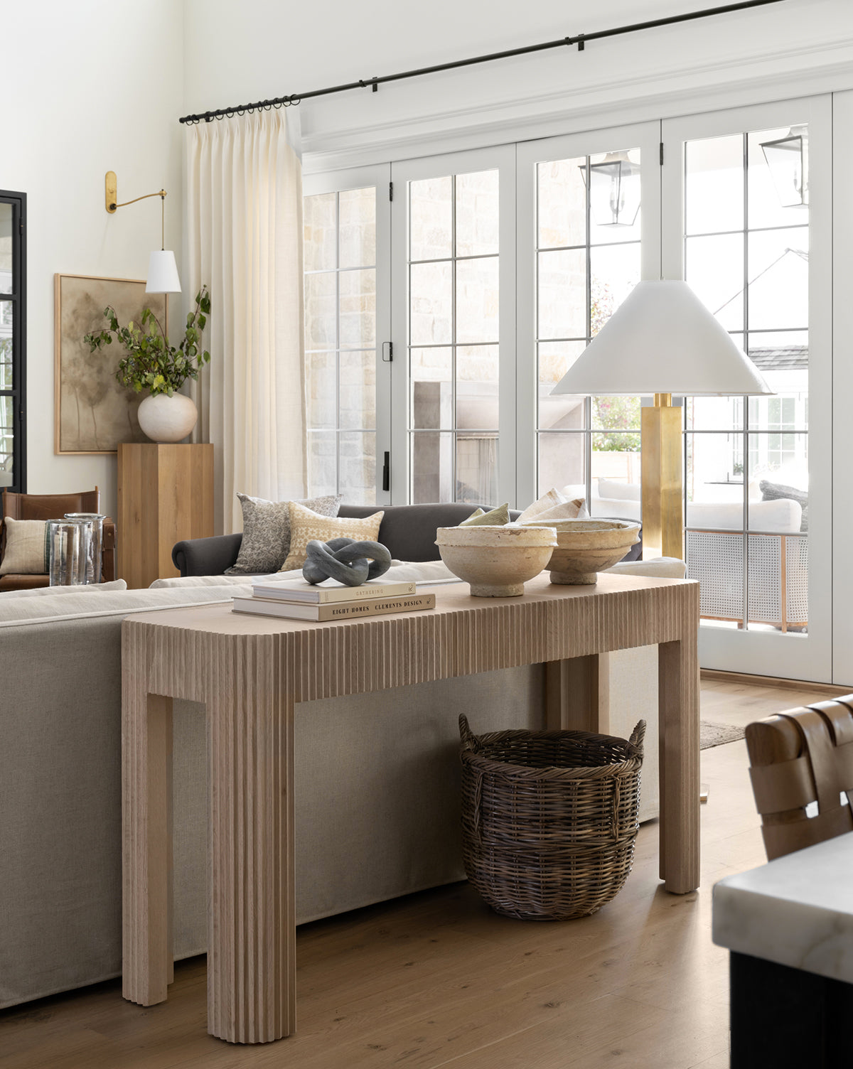 A bright, modern living room has a light wood console table behind a beige sofa, topped with bowls and books. Large windows with white curtains let in natural light. The Mckell Basket sits under the table for extra storage.