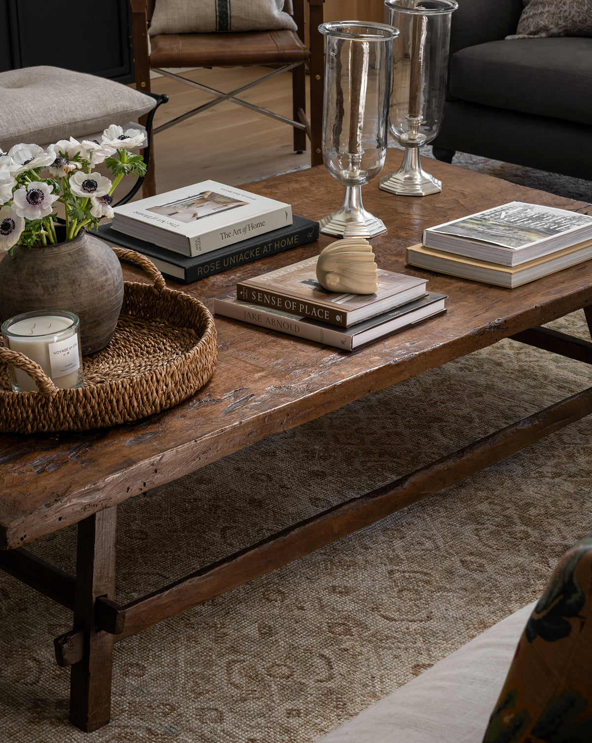 A rustic wooden coffee table styled with books, candles, glass hurricanes, white anemones in a vase, and a woven tray sits on a patterned rug next to the Baroque Shell Object, creating a cozy and elegant living room scene.