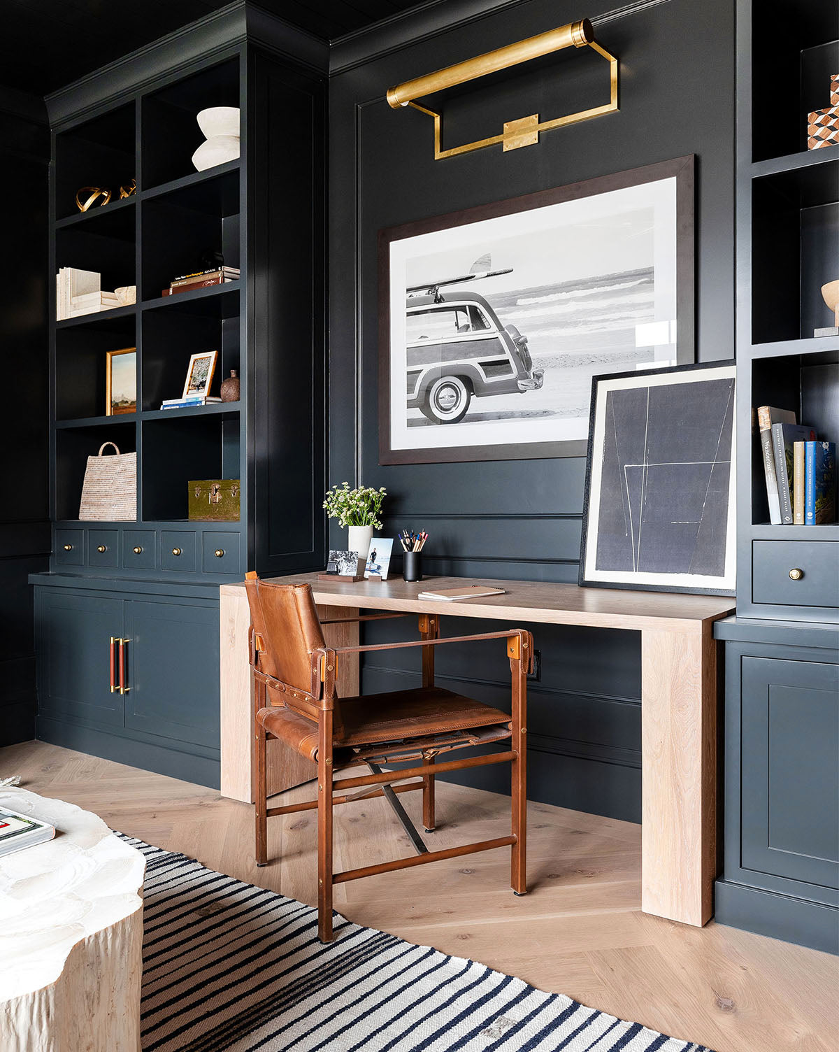 A modern home office features dark built-in shelves, a light wood desk, a brown leather chair, and black and white wall art called Gone Surfin depicting a vintage car. A striped rug brings cozy texture to the stylish space.