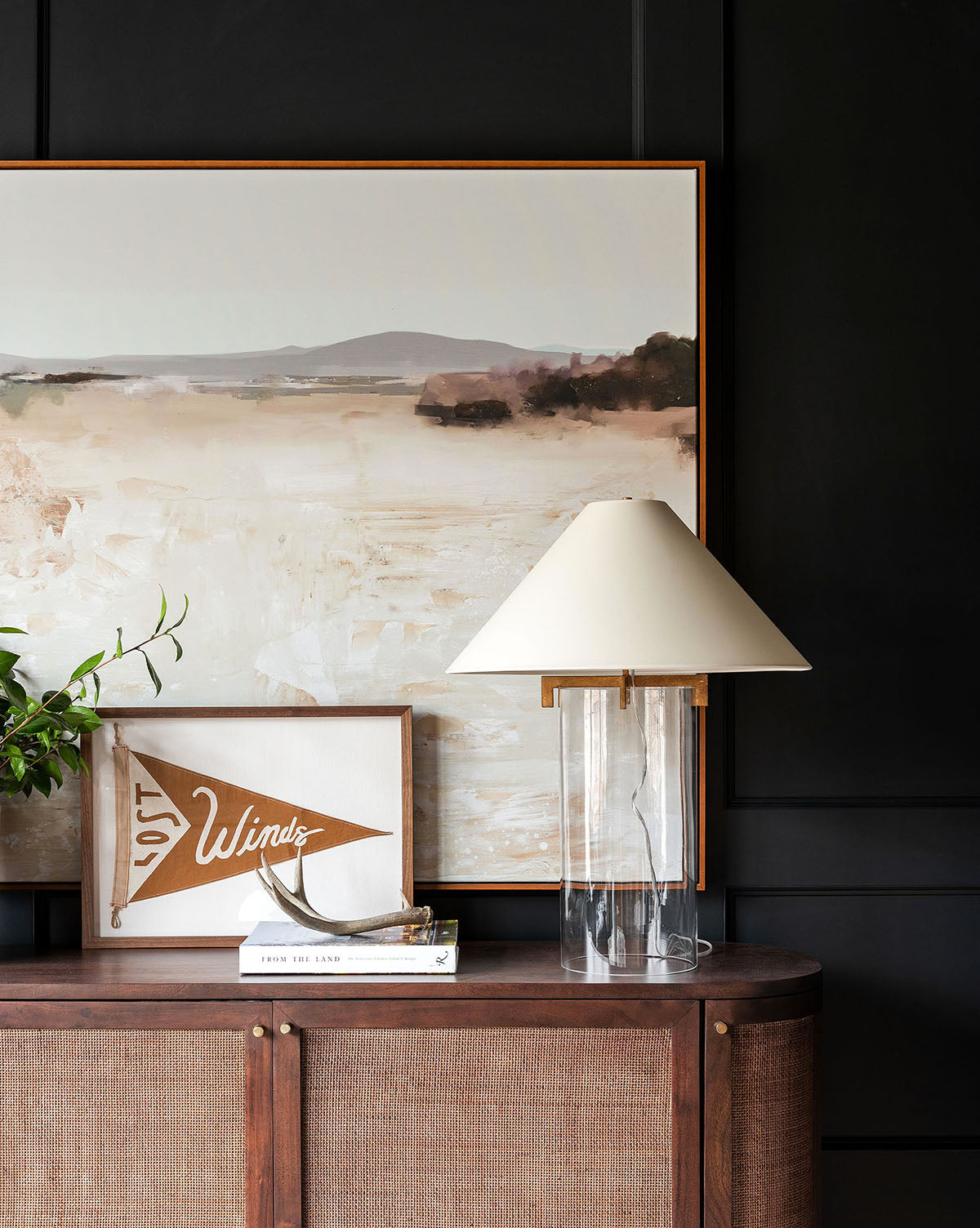A modern sideboard with books, the Brooks Table Lamp in crystal white, a framed WINDS pennant, and a muted earth-toned landscape painting stands against a dark paneled wall, by McGee & Co.