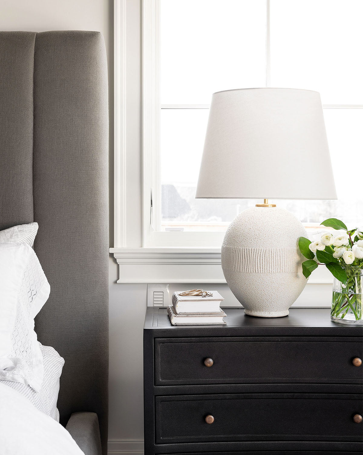 A bedside table with a natural texture features the Toulon Table Lamp by Visual Comfort & Co. Signature with a linen shade, alongside a vase of white flowers, books, and glasses next to a gray upholstered bed in a modern bedroom. McGee & Co.