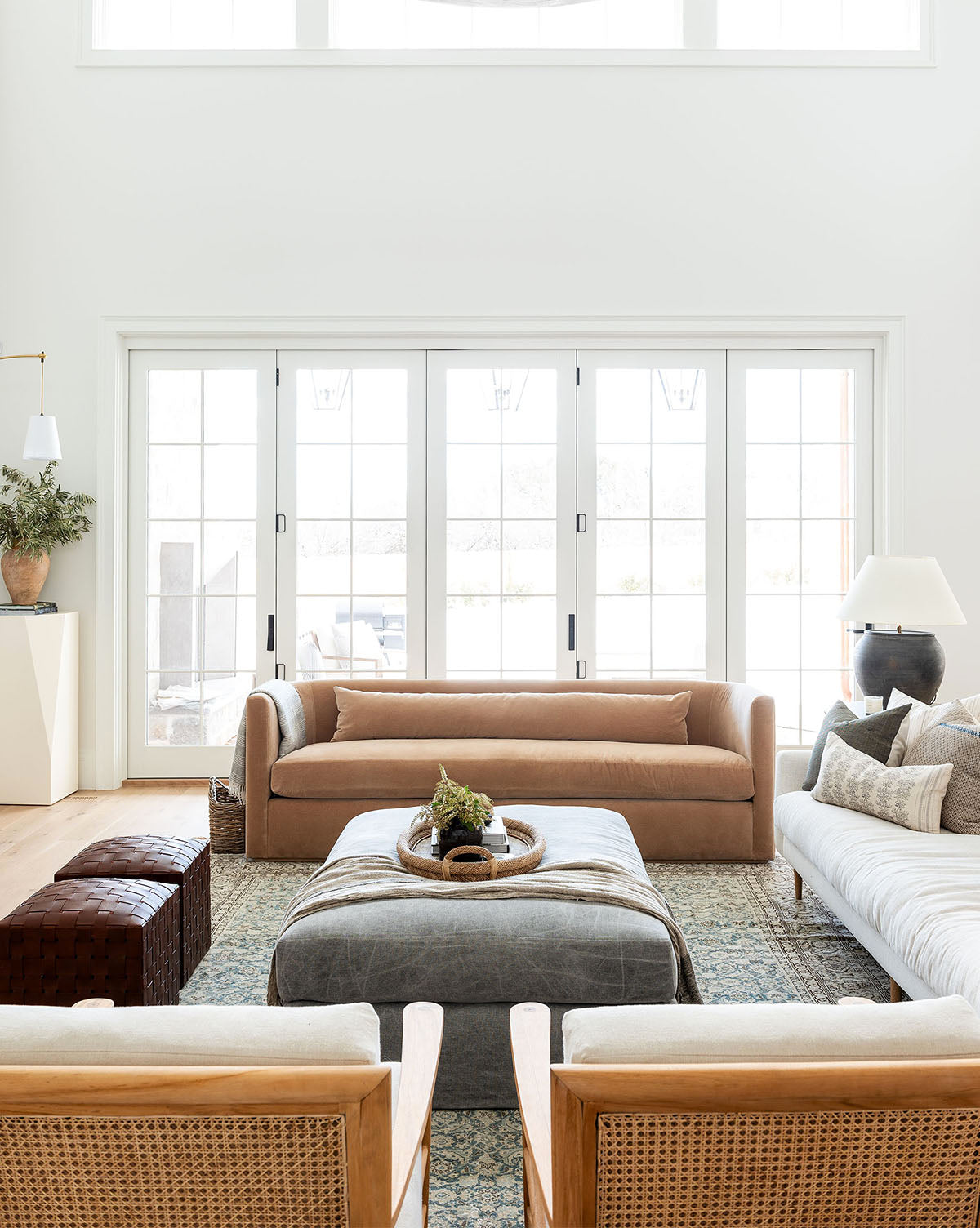 A bright, modern living room with large windows features a tan Reese Sofa (84-96), a white sofa with down-wrapped cushions, two handcrafted alder wood armchairs, a gray ottoman as a coffee table, and neutral decor such as cushions, a lamp, and a potted plant by McGee & Co.