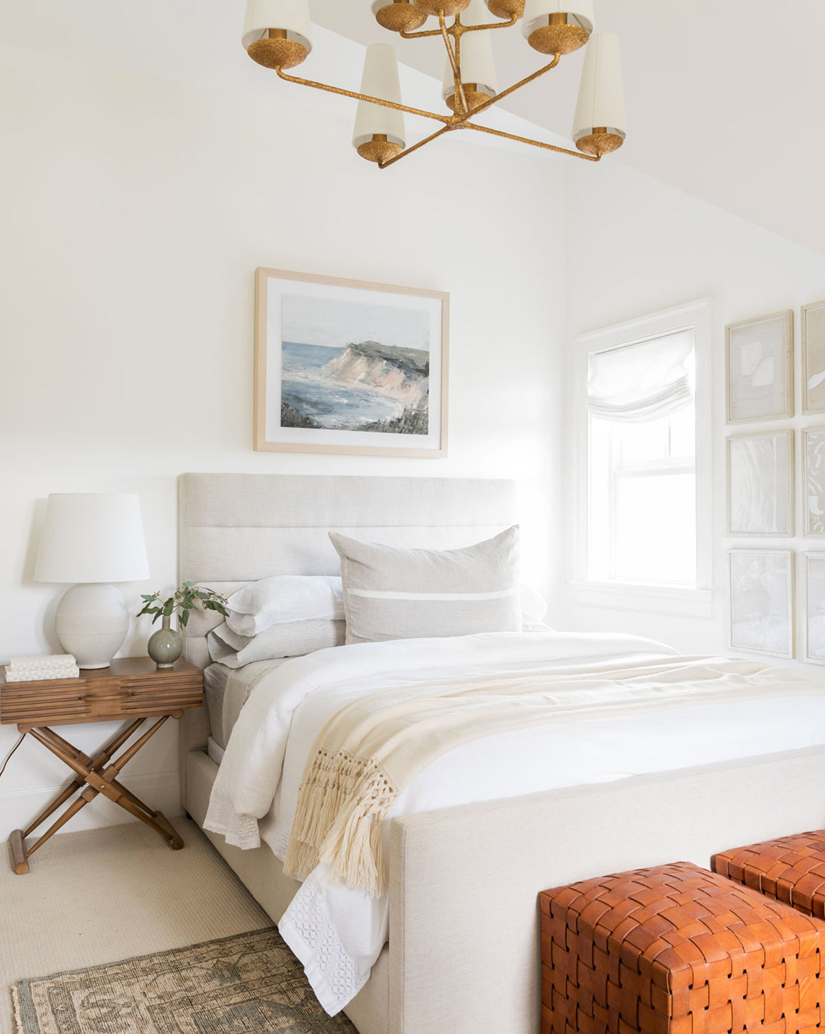 A bright, airy bedroom with the Lila Bed in a beige quilted frame, white bedding, and neutral pillows. A wooden side table with lamp and plant sits beside it. Art is displayed above, with a gold ceiling light fixture completing the space.