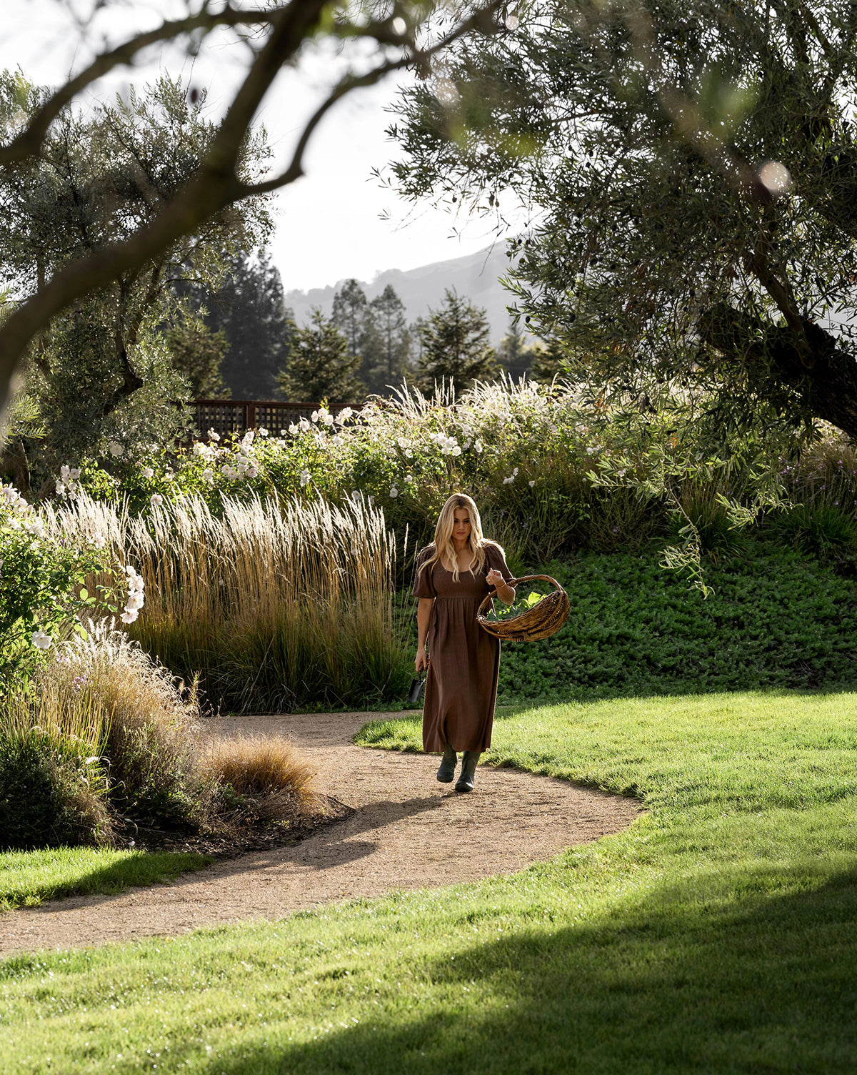 A woman in a long brown dress walks through a garden path as the air fills with the aromatic herbal and citrus scent of Pura x Studio McGee Neroli Basil Home Fragrance Refill.