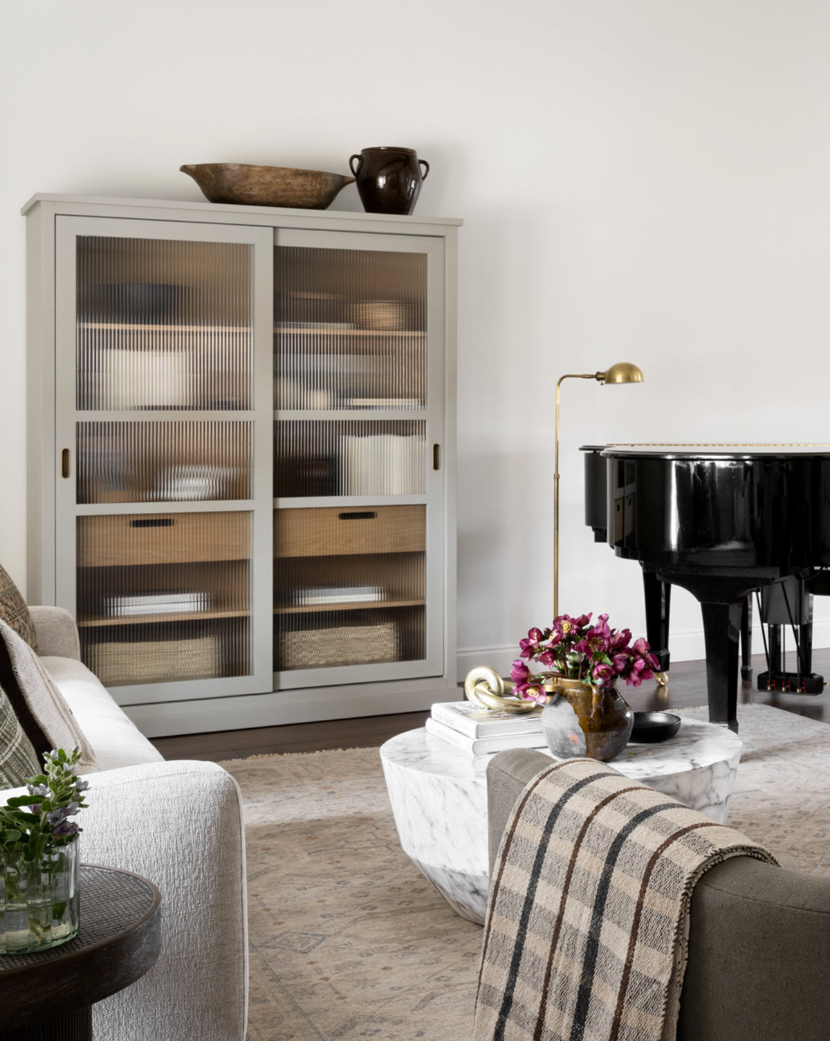 A cozy living room with a white cabinet of baskets, a marble coffee table with books and flowers, a beige sofa with a plaid throw, the Old Pharmacy Floor Lamp for adjustable height lighting, and a black grand piano against a white wall, by McGee & Co.