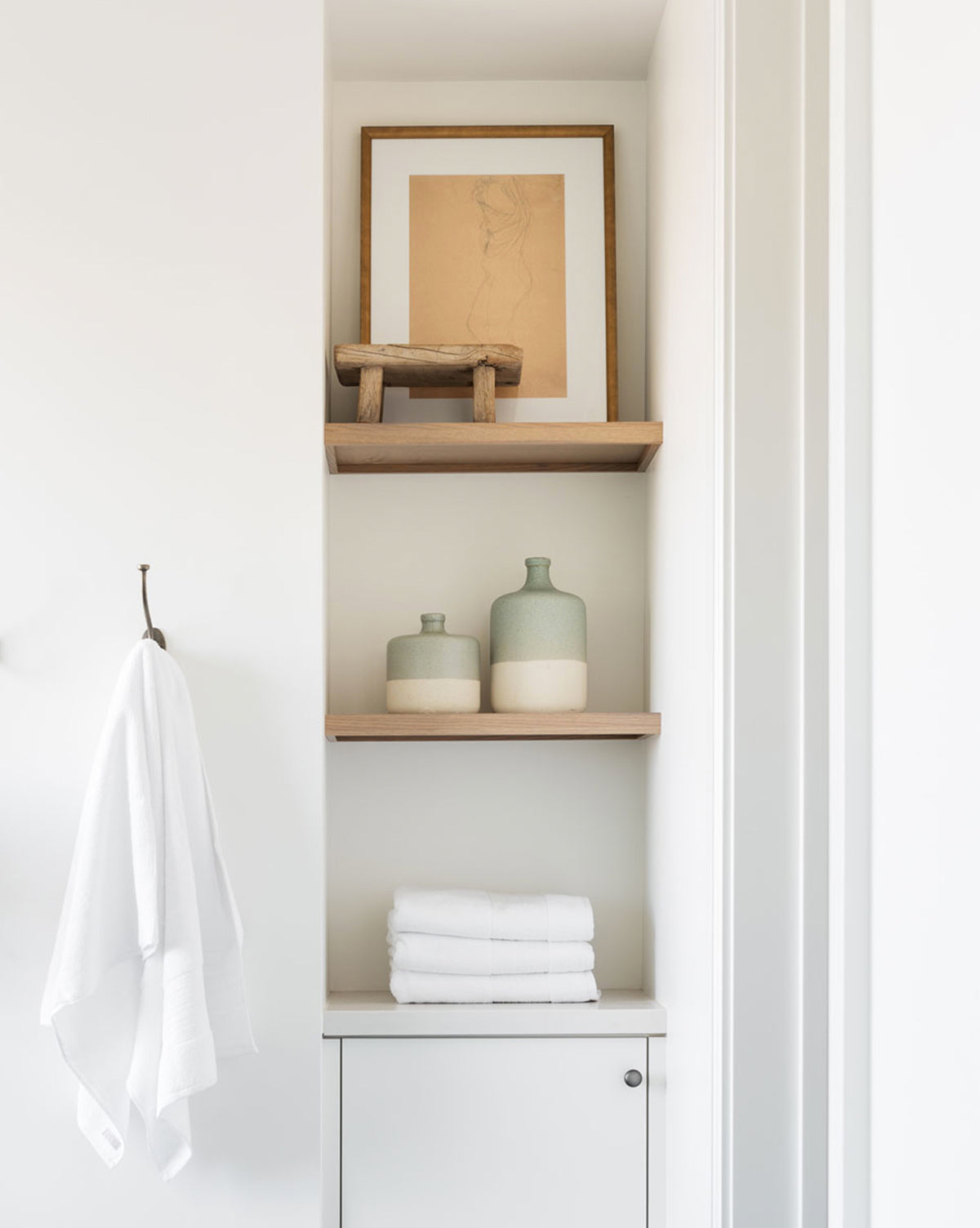 A minimalist bathroom nook features two wooden shelves with green and cream ceramic bottles, a small stool, folded white towels, and framed art in progress. The display is completed by the Sketched Figure artwork and a towel on a simple frame.
