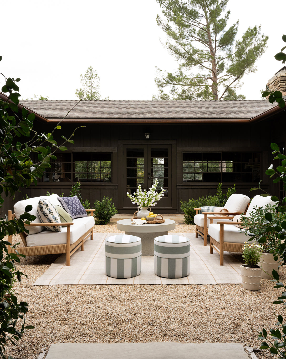 A cozy outdoor patio featuring cushioned chairs, a weather-resistant Makerspalm Haviland Striped Ottoman, and a round coffee table on a rug amid plants and gravel, set before a dark wooden house with large windows and trees. McGee & Co.