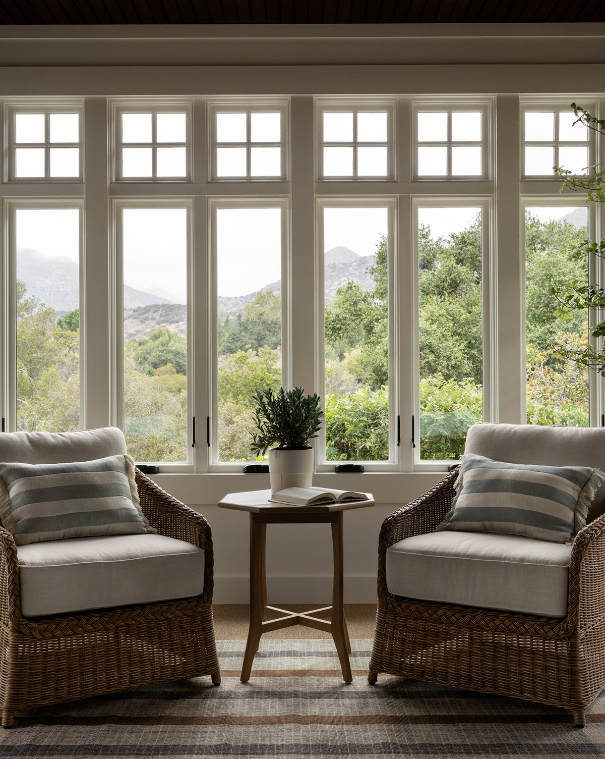 Two Haviland Outdoor Lounge Chairs with striped cushions face each other by a small round table with a plant and book, set in front of large windows with views of trees and distant mountains, by McGee & Co.
