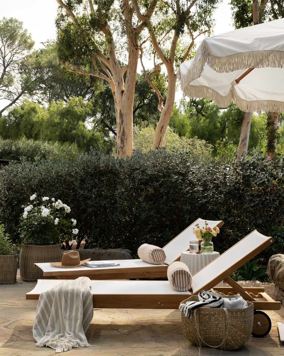 Two Cadie Outdoor Chaise lounge chairs with white, weather-resistant cushions sit side by side outdoors under a fringed umbrella, accented by striped pillows, a hat, blanket, drinks, and a basket amid lush garden greenery by McGee & Co.