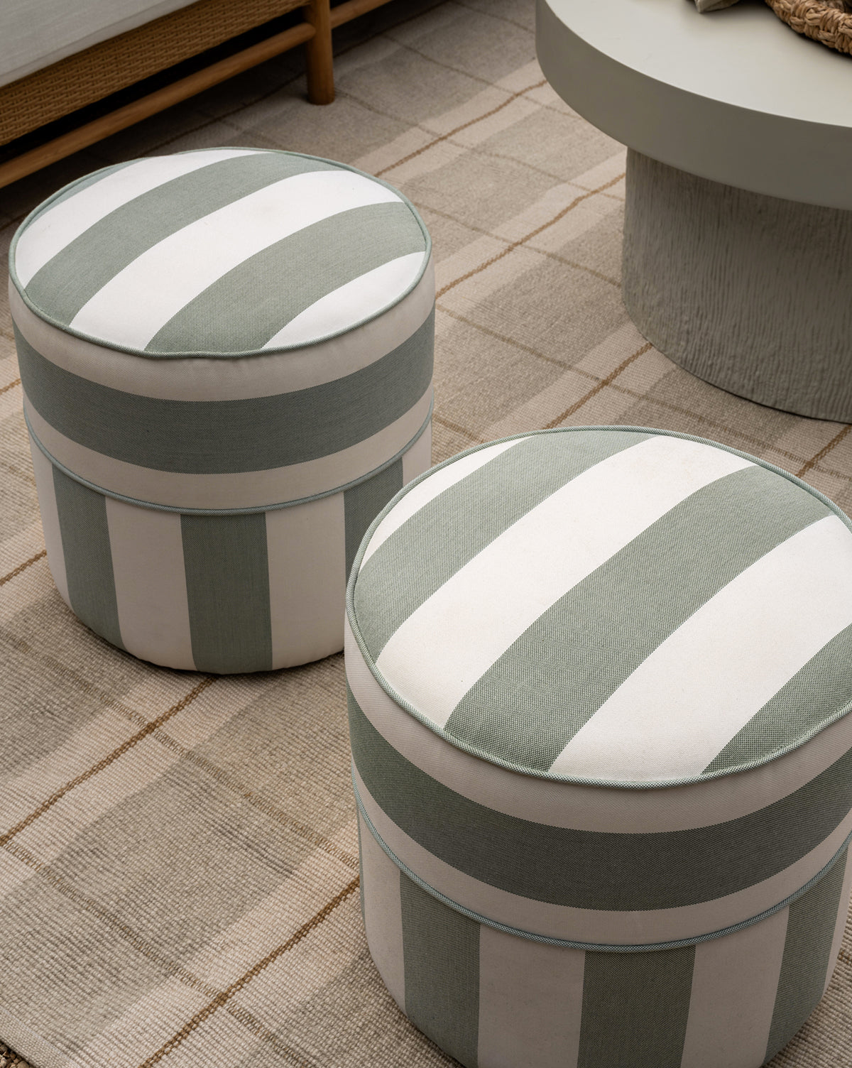 Two Makerspalm Haviland Striped Ottomans with vertical green and white stripes, made for outdoor use, rest on a beige checkered rug in a modern living room next to a round light-gray table. McGee & Co.