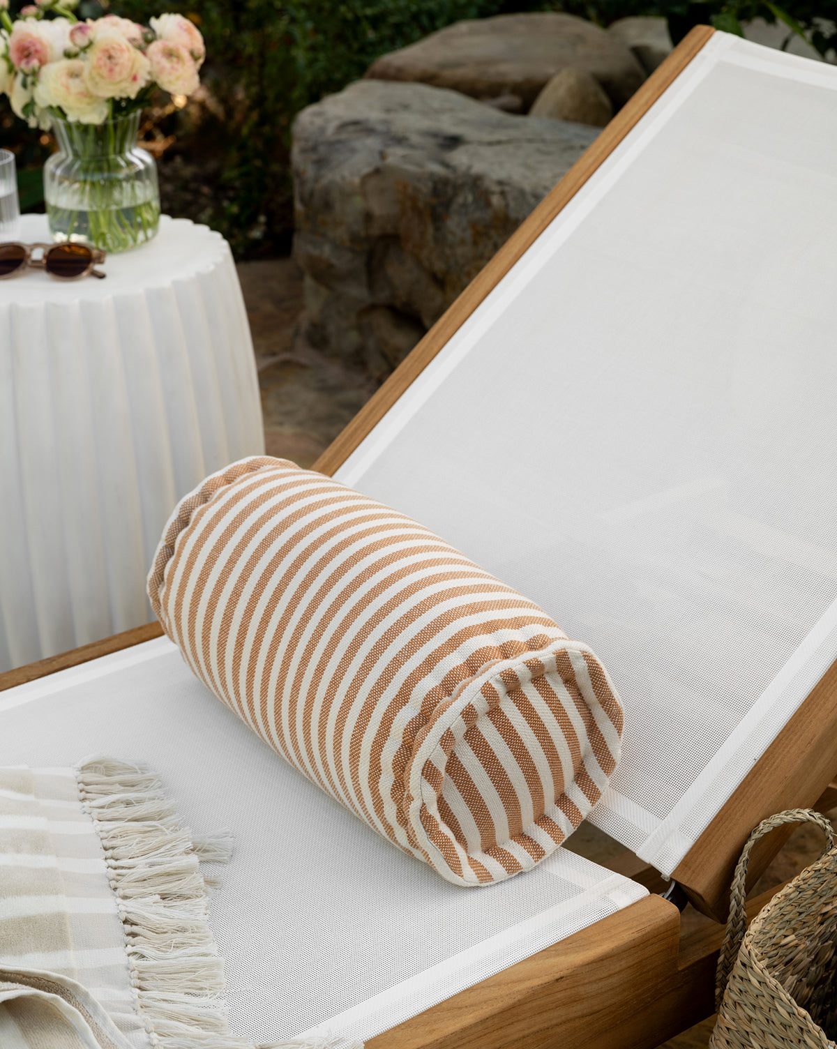 A white lounge chair with a weather-resistant pillow, the Striped Indoor/Outdoor Bolster Pillow in tan and white, and a fringed throw sits by a round table with flowers and sunglasses. Stones and greenery add to the outdoor decor backdrop, by McGee & Co.