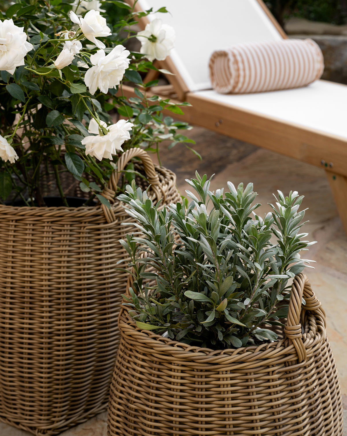 Two Lightwash Handled Planter Baskets with green indoor plants—one blooming white flowers—are placed outdoors beside a wooden lounge chair featuring a white cushion and striped bolster pillow, by McGee & Co.