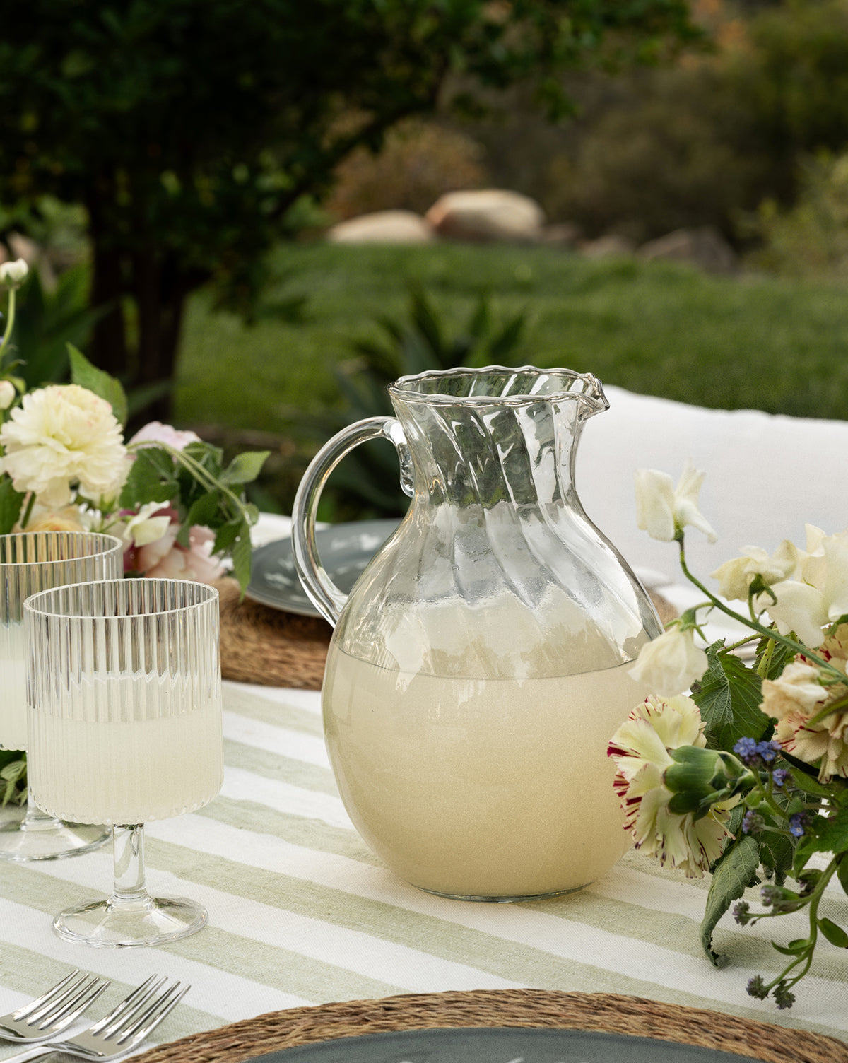 The Adderley Pitcher, made of mouth-blown glass and filled with a light beverage, sits on an outdoor table alongside two matching glasses, floral arrangements, and greenery in the background, by McGee & Co.