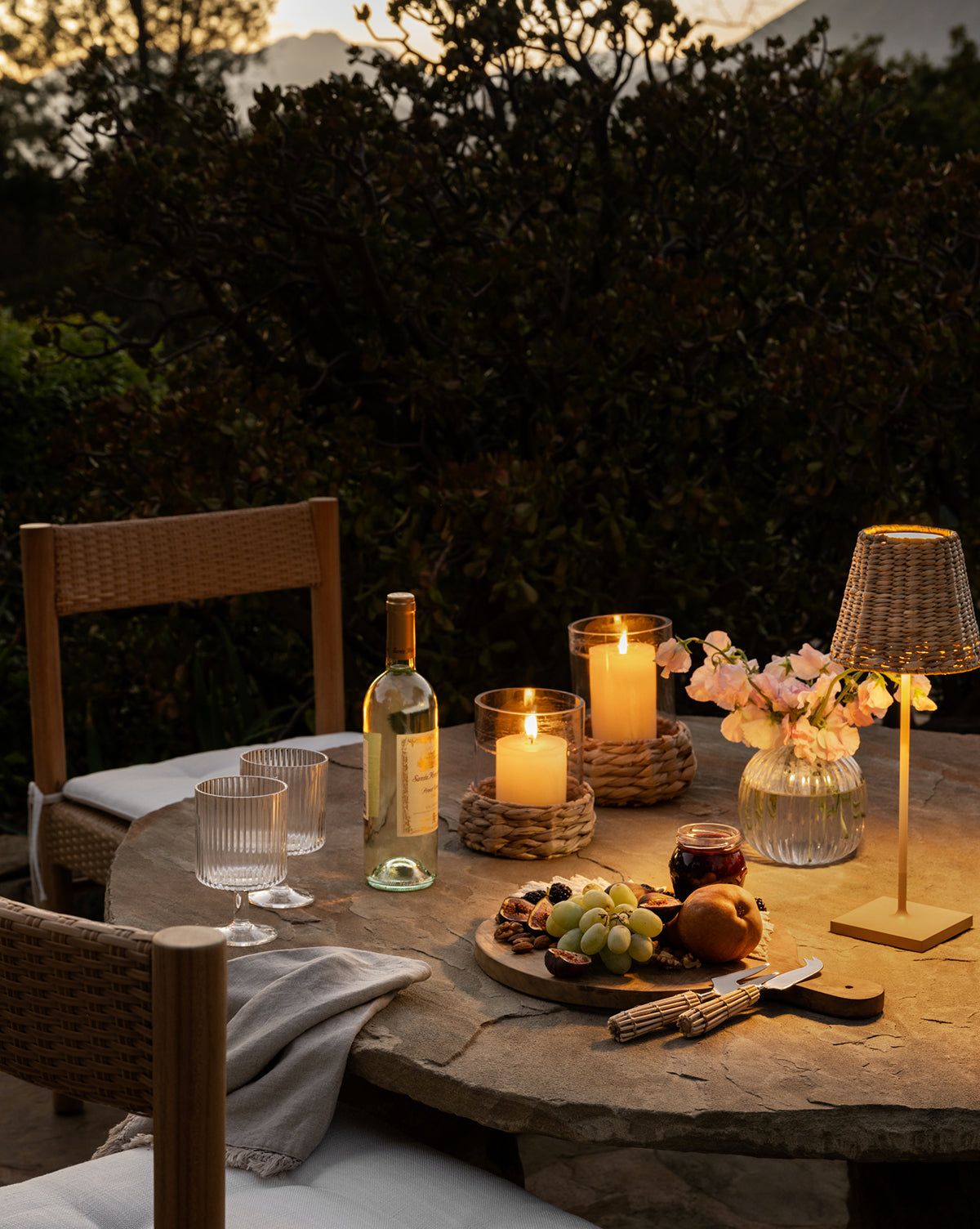 At dusk, a round stone table for two features wine, grapes, cheese, and bread. Woven Hurricane candle holders and a small lamp cast warm light on the scene with wicker chairs and natural accents.