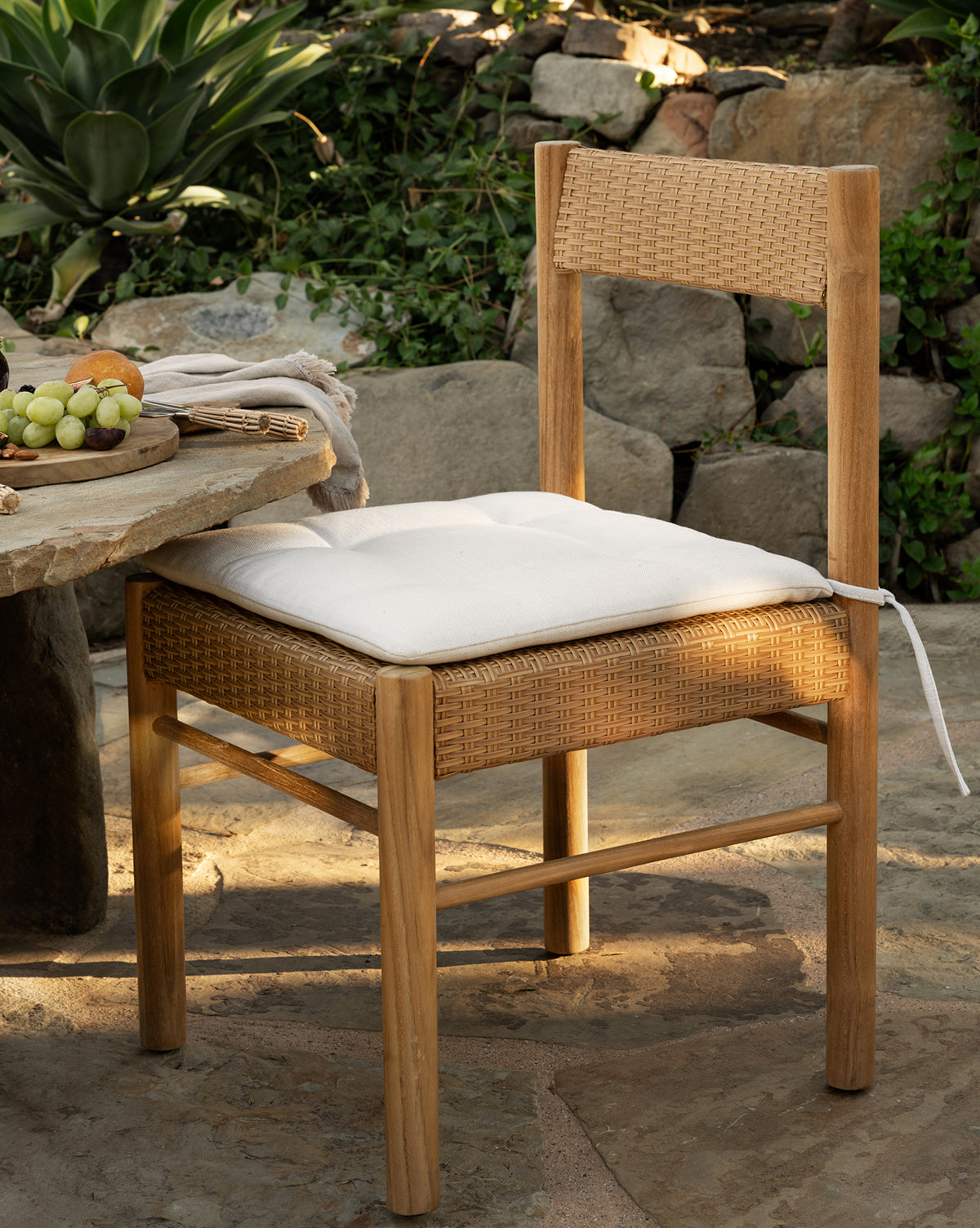 The Simeon Outdoor Dining Chair, featuring a woven rattan seat and backrest with a white cushion, sits by a stone table holding grapes in a garden surrounded by rocks and lush green plants, by McGee & Co.