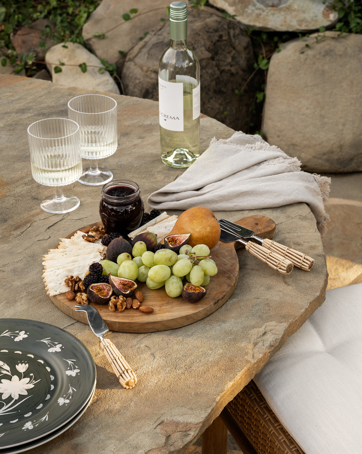 A rustic outdoor table set features the Boylan Bread Board topped with green grapes, figs, pear, walnuts, cheese, and jam. Nearby are two glasses, a bottle of white wine, and a folded beige napkin on the stone surface.