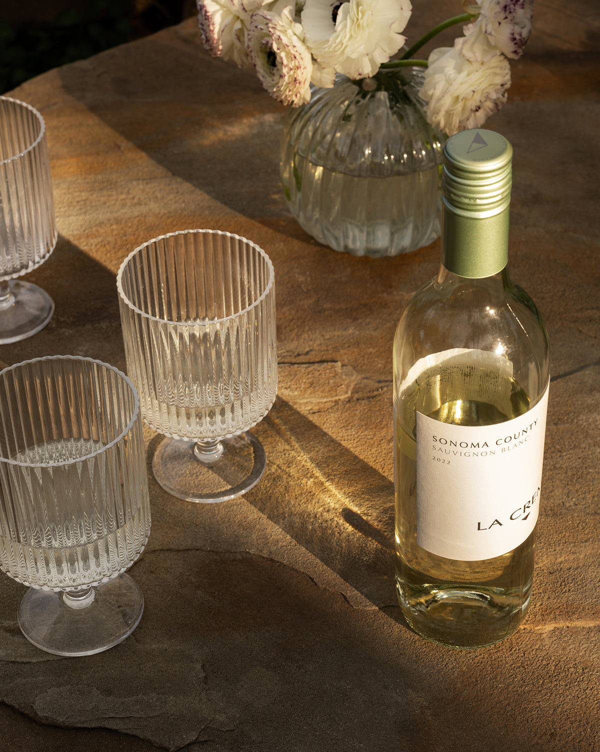 A bottle of Sonoma County Sauvignon Blanc sits on a stone table with Sanibel Acrylic Goblets (Set of 4) and a vase of white flowers, creating elegant outdoor table settings in soft, warm sunlight.