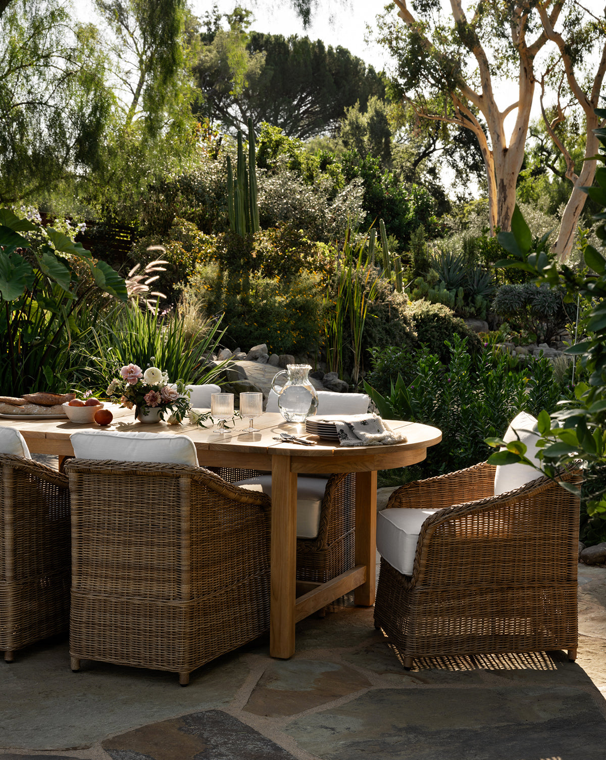A round wooden table is set for a meal with Haviland Outdoor Dining Chairs featuring weather-resistant wicker and white cushions. The table has glassware, plates, napkins, and a floral centerpiece, surrounded by lush green patio plants by McGee & Co.