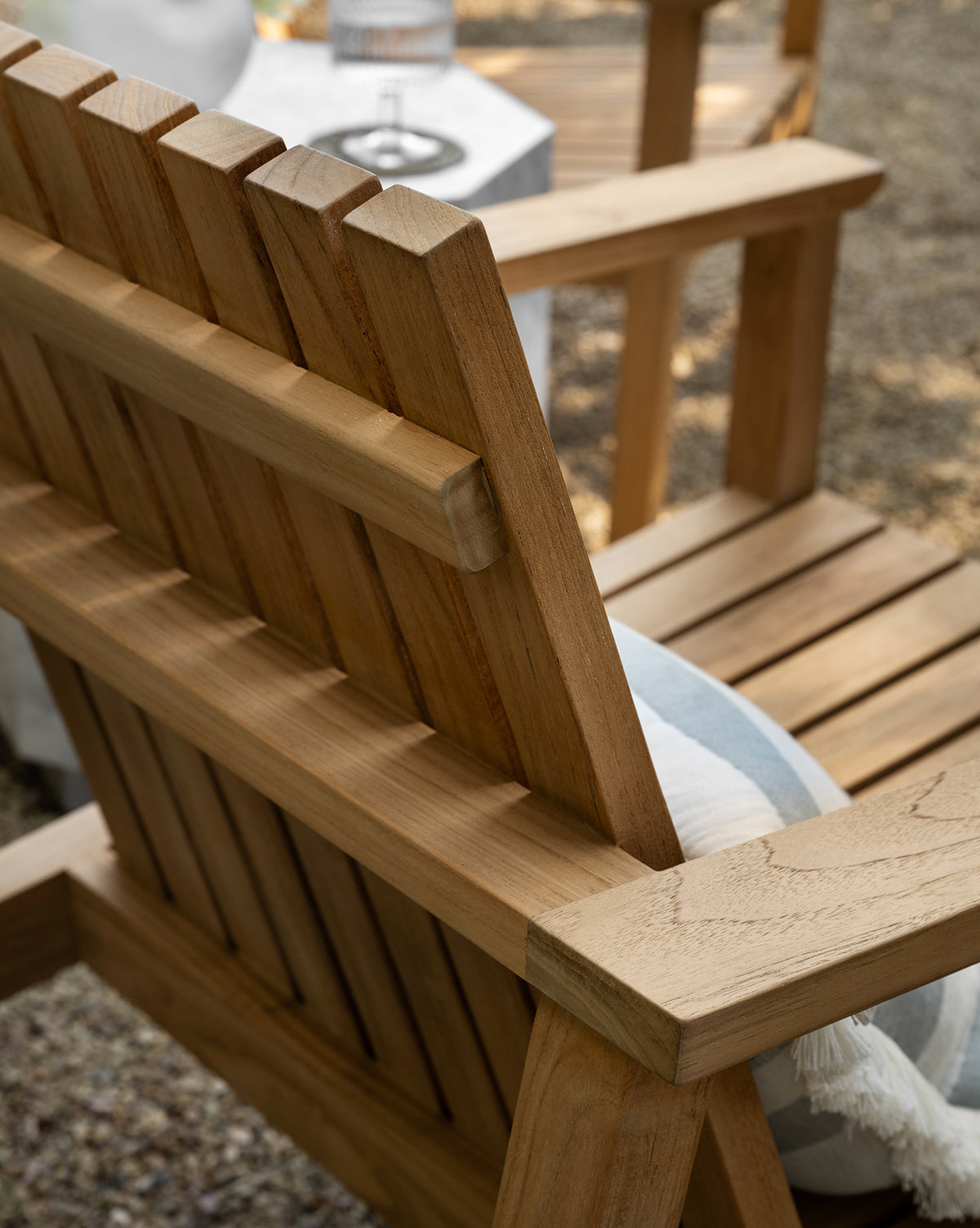 Close-up of the Gilda Outdoor Lounge Chair in teak wood, featuring a slatted back and seat with a blue and white striped cushion. In the background, a small table holds a glass and carafe on a gravel surface, by McGee & Co.