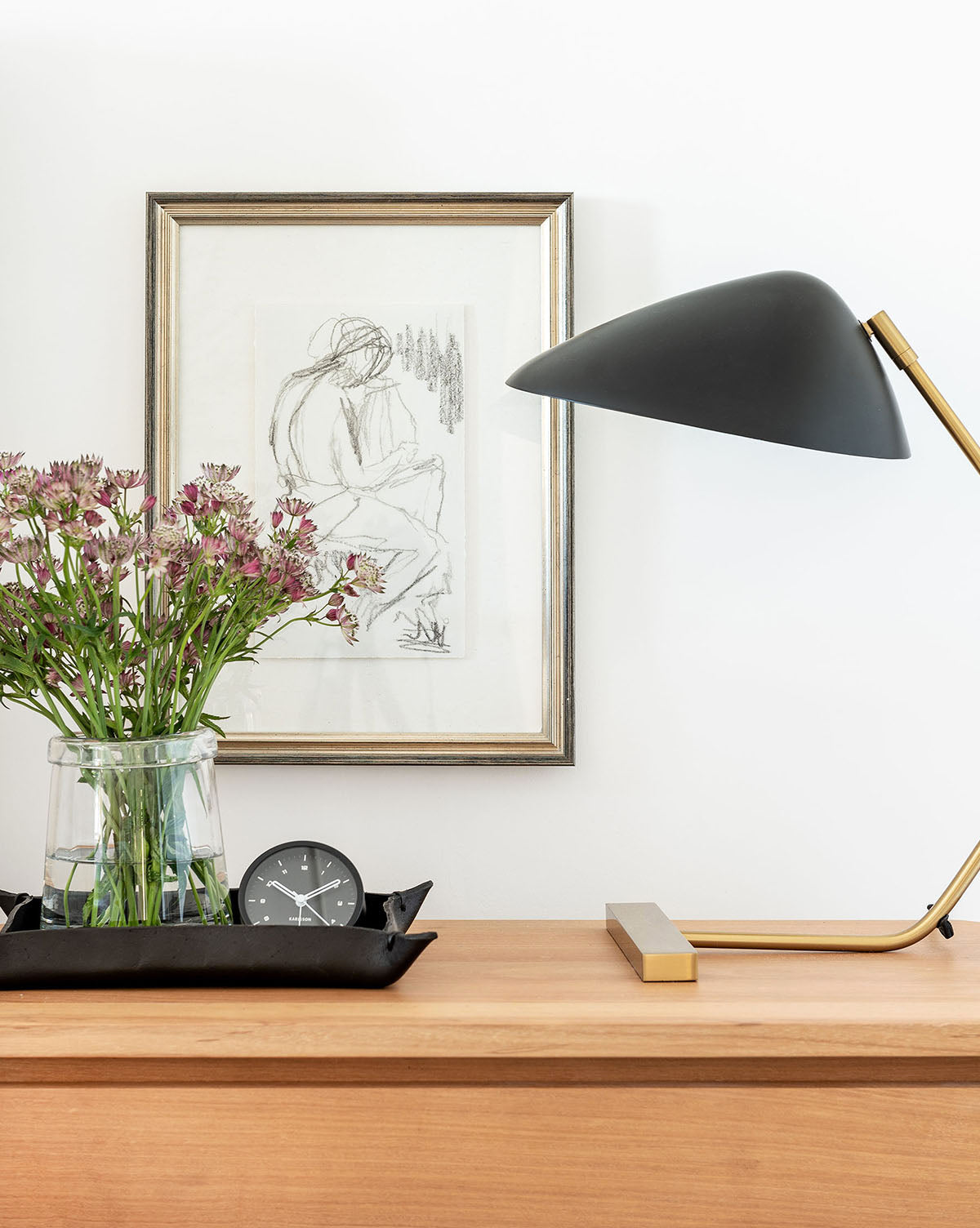 A modern wooden desk with a black table lamp, purple flowers in a vase, a small black clock, and the Figure Sketch framed as gallery wall decor on the white wall behind.