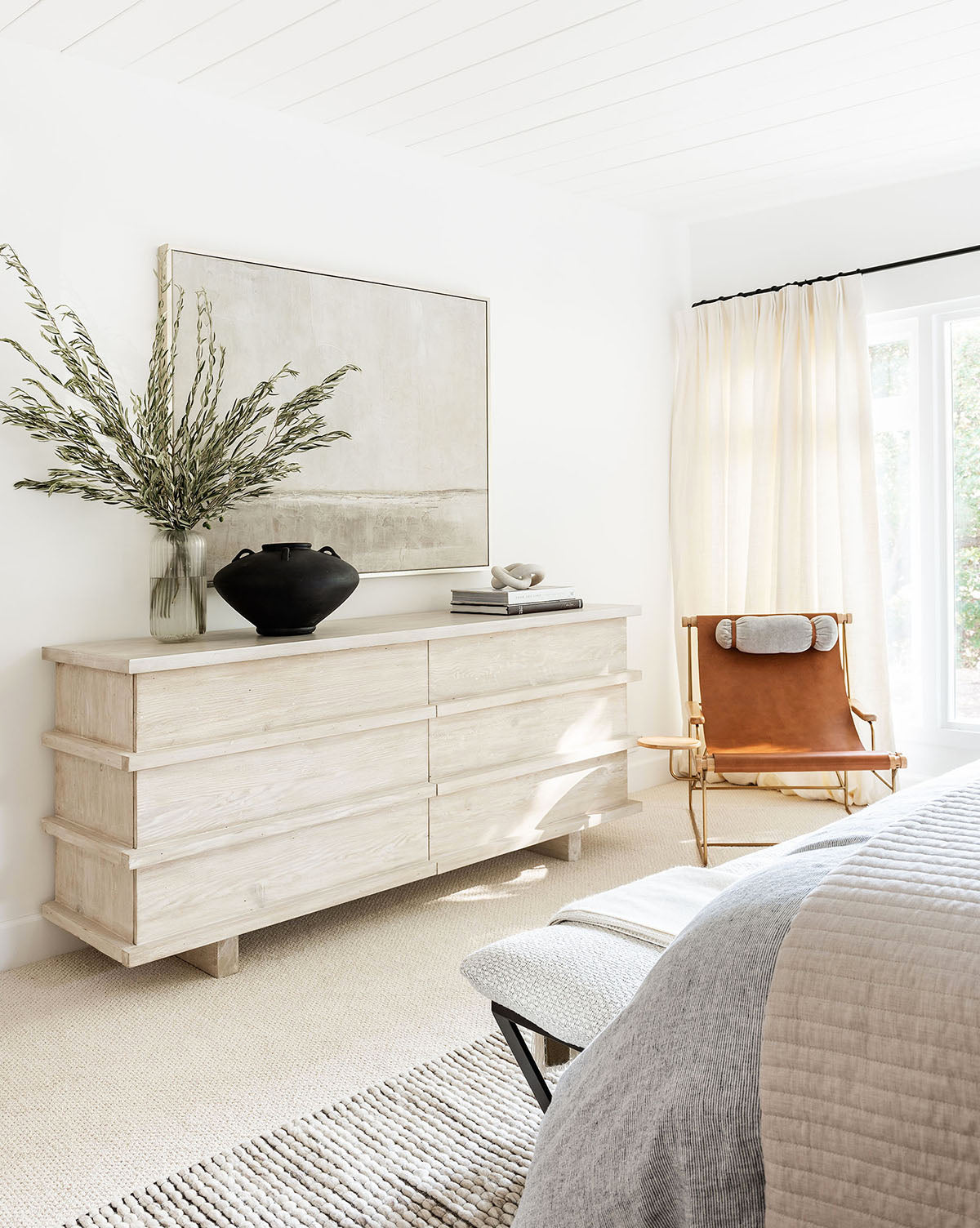 A bright, minimalist bedroom features the Gulf Shores dresser in light wood, serene gray and green decor, a large abstract painting, a black vase with greenery, a tan leather chair, and cream curtains welcoming natural light.
