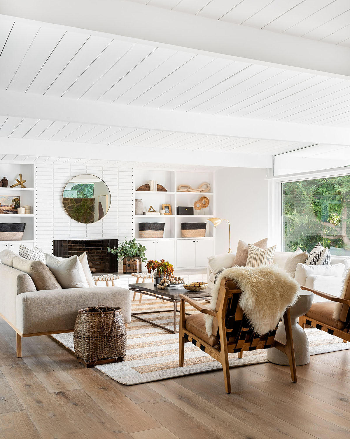A bright, modern living room with neutral furniture, wood accents like an ash wood frame Lanston accent chair with leather cushions, woven decor, a striped rug, and a fireplace. Large windows let in lots of natural light and show greenery outside.