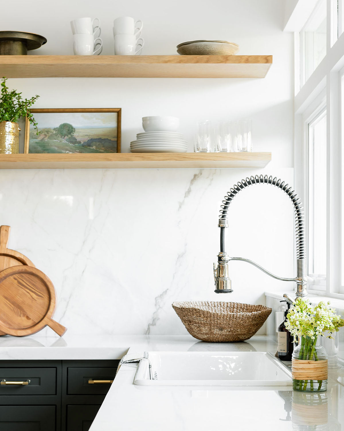 A modern kitchen featuring the Backroads white marble backsplash, open wooden shelves with dishes and decor, a deep sink with spring faucet, woven basket, and fresh flowers on the counter brings vintage charm to the inviting space.