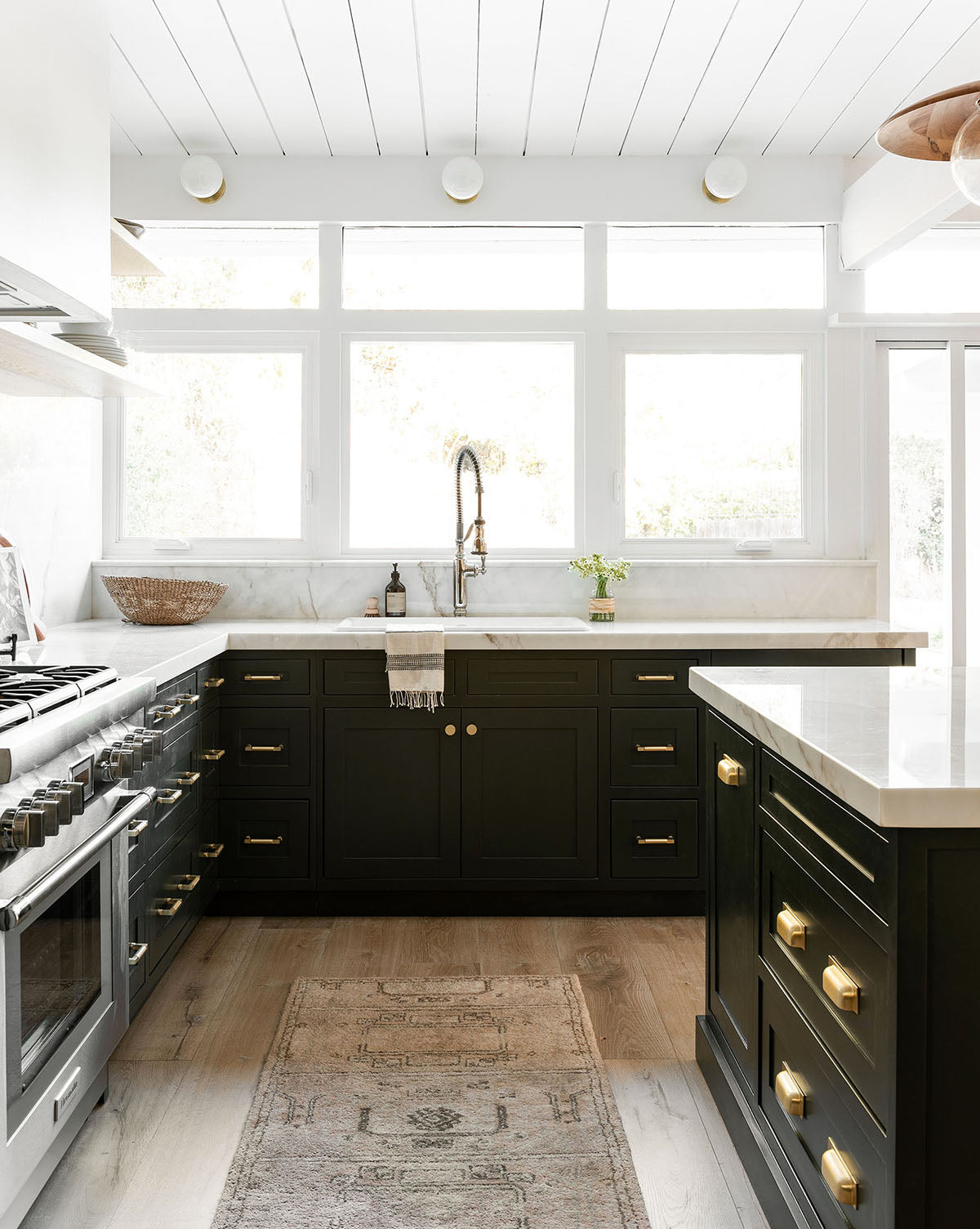 Bright modern kitchen with black cabinets, gold hardware, white countertops, stainless steel appliances, a farmhouse sink, large windows, and an Ellington Fawn Hand-Tufted Wool Rug on light wood floors, by McGee & Co.
