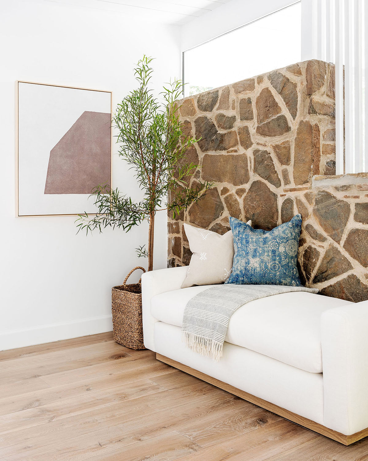 A cozy living space features a white sofa with blue and beige pillows, a gray throw, a stone accent wall, light wood flooring, a basket, a tall potted plant, and curated wall art framed as the Entering Shape 2 abstract piece.