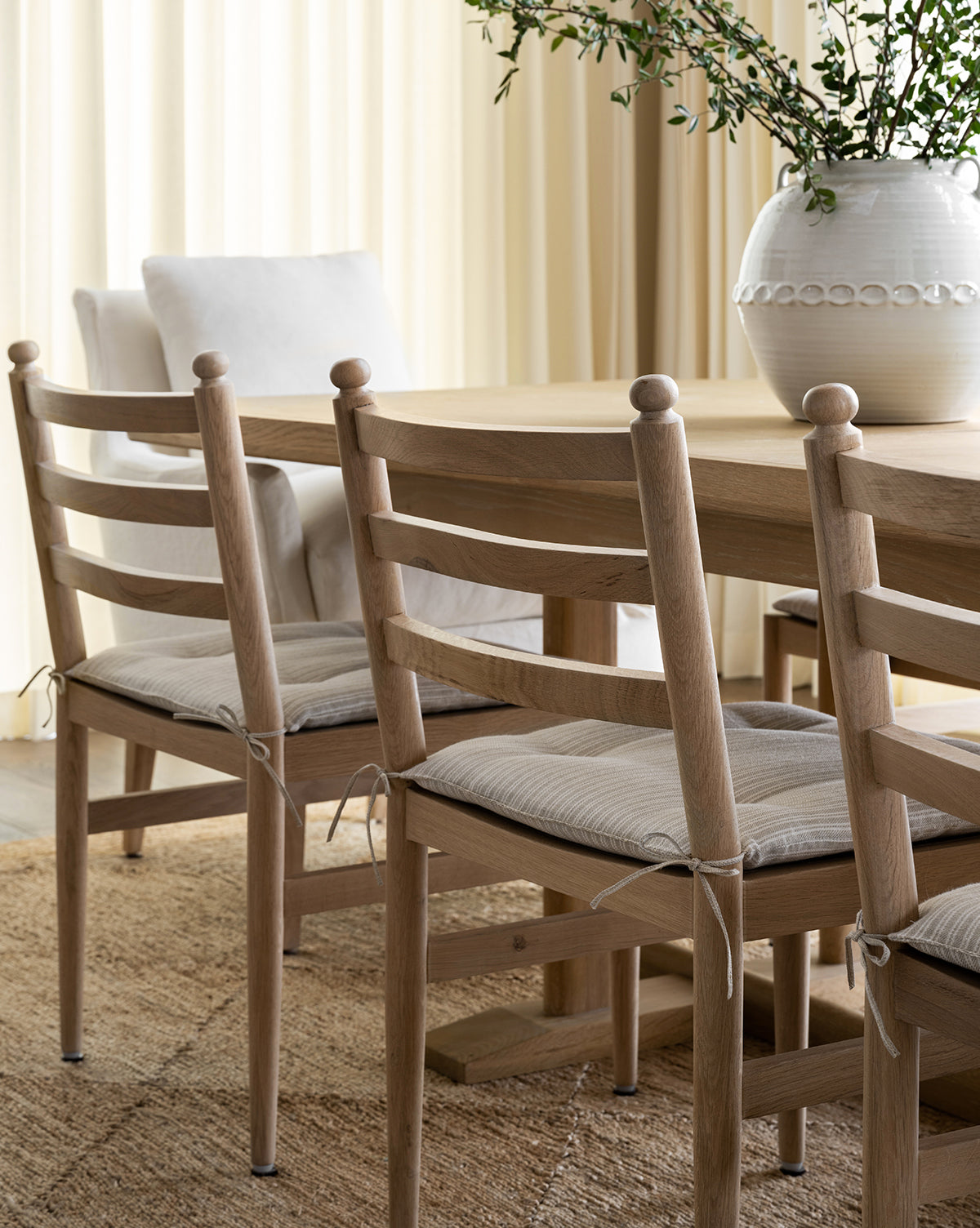 A light white oak dining table with Romany Dining Chairs featuring beige seat cushions and a woven rug. A large white vase with greenery decorates the table, set in a room with soft, neutral drapes, by McGee & Co.