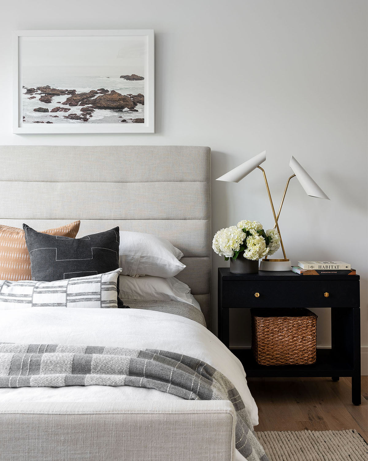 A modern bedroom featuring the Lila Bed with a quilted frame, layered pillows, a gray and white blanket, a black nightstand with books, a basket of white flowers, a double-headed lamp, and a framed photo of rocks and water above.