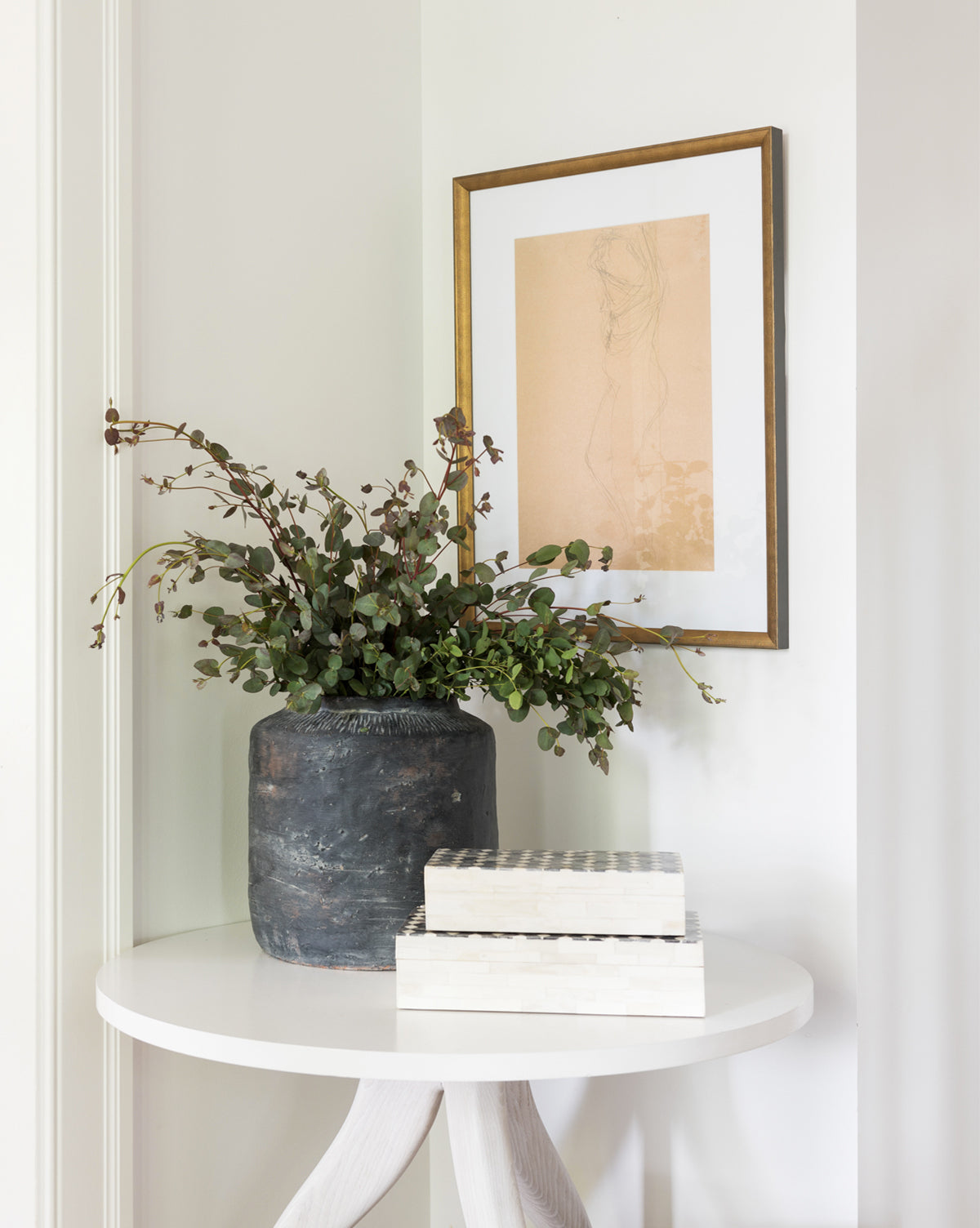 A round white table displays a dark ceramic vase with green branches and two stacked boxes. Above, the Sketched Figure artwork in a simple frame brings an artistic touch to the white wall.