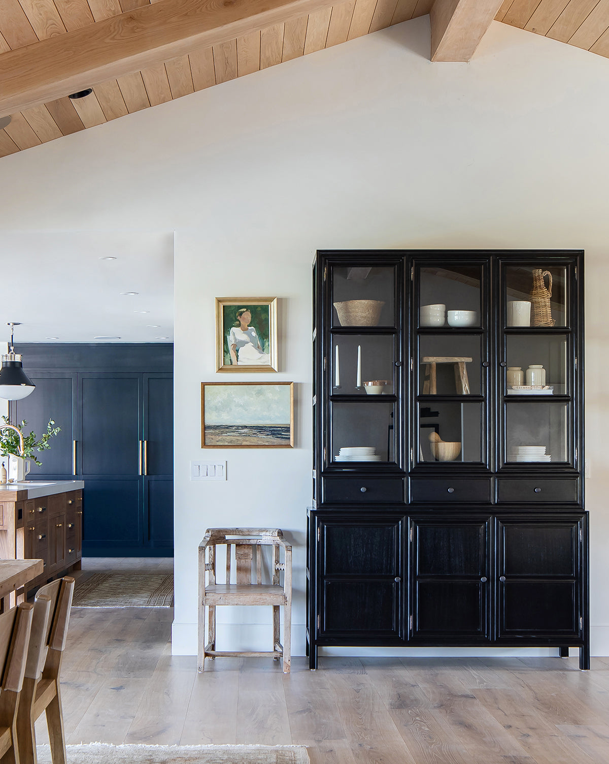 A modern dining area with wood floors, vaulted ceiling, black glass-front cabinet, small wooden stool, and two framed Ponder in the Park 1 abstract art pieces on the wall, looking into a kitchen with dark cabinets.