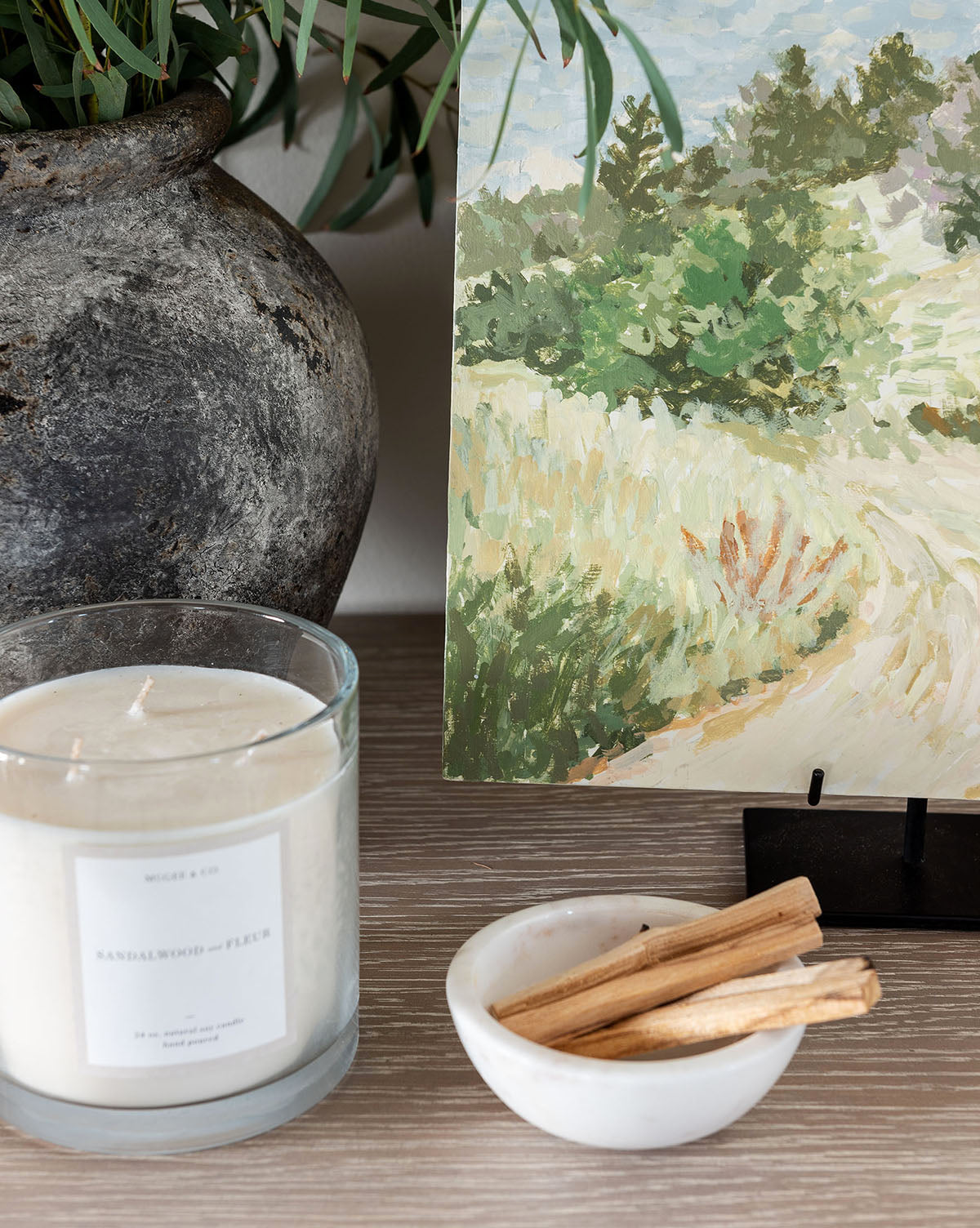 A white candle in a glass jar, a small Marble Nesting Bowl from the set of 3 with wood sticks, and a potted plant sit on a wooden surface beside a landscape painting featuring greenery and a path.