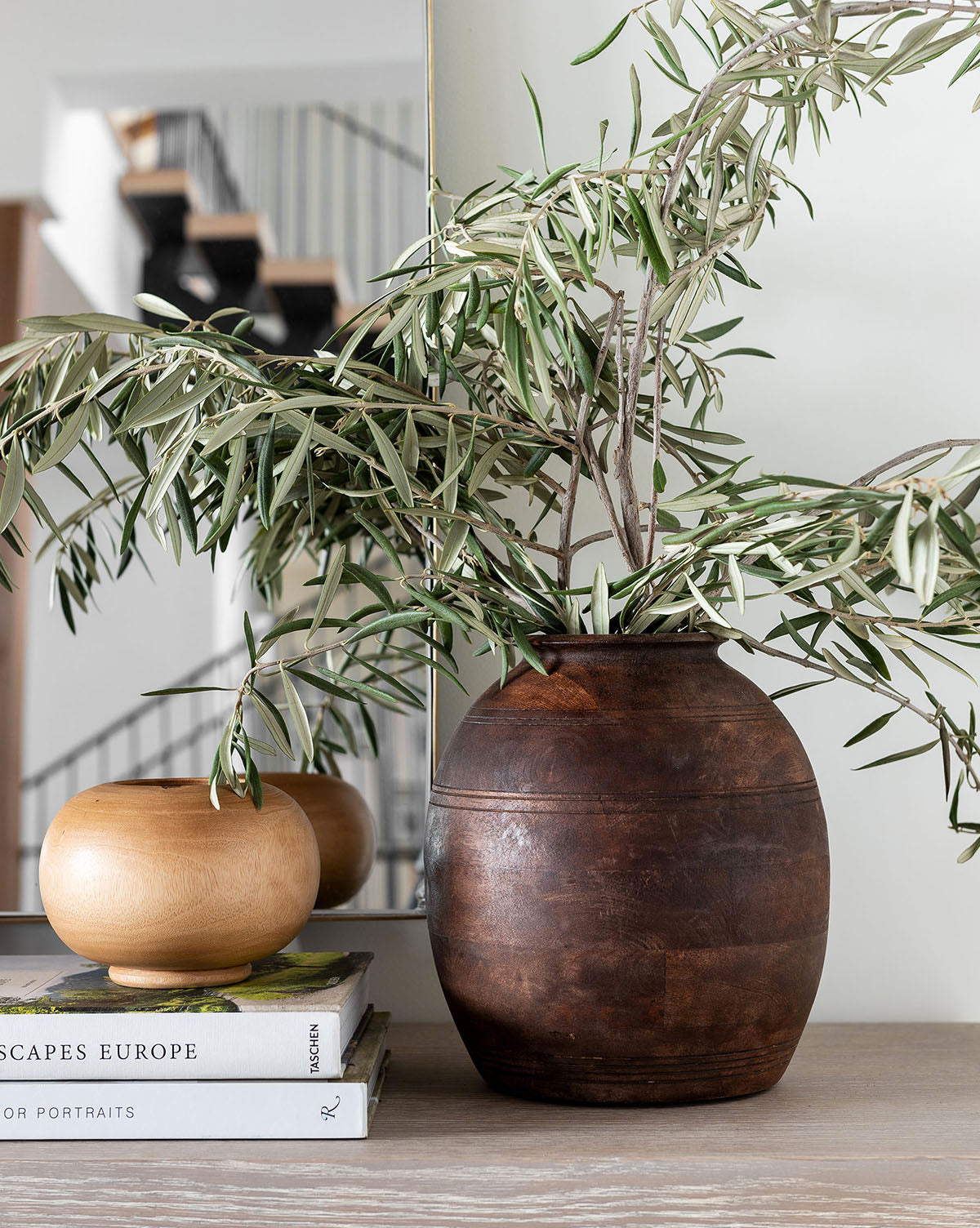 The Aged Wood Vase with leafy branches is displayed on a light wooden table next to a small bowl and stacked books, all styled beautifully with a mirror and staircase in the background.