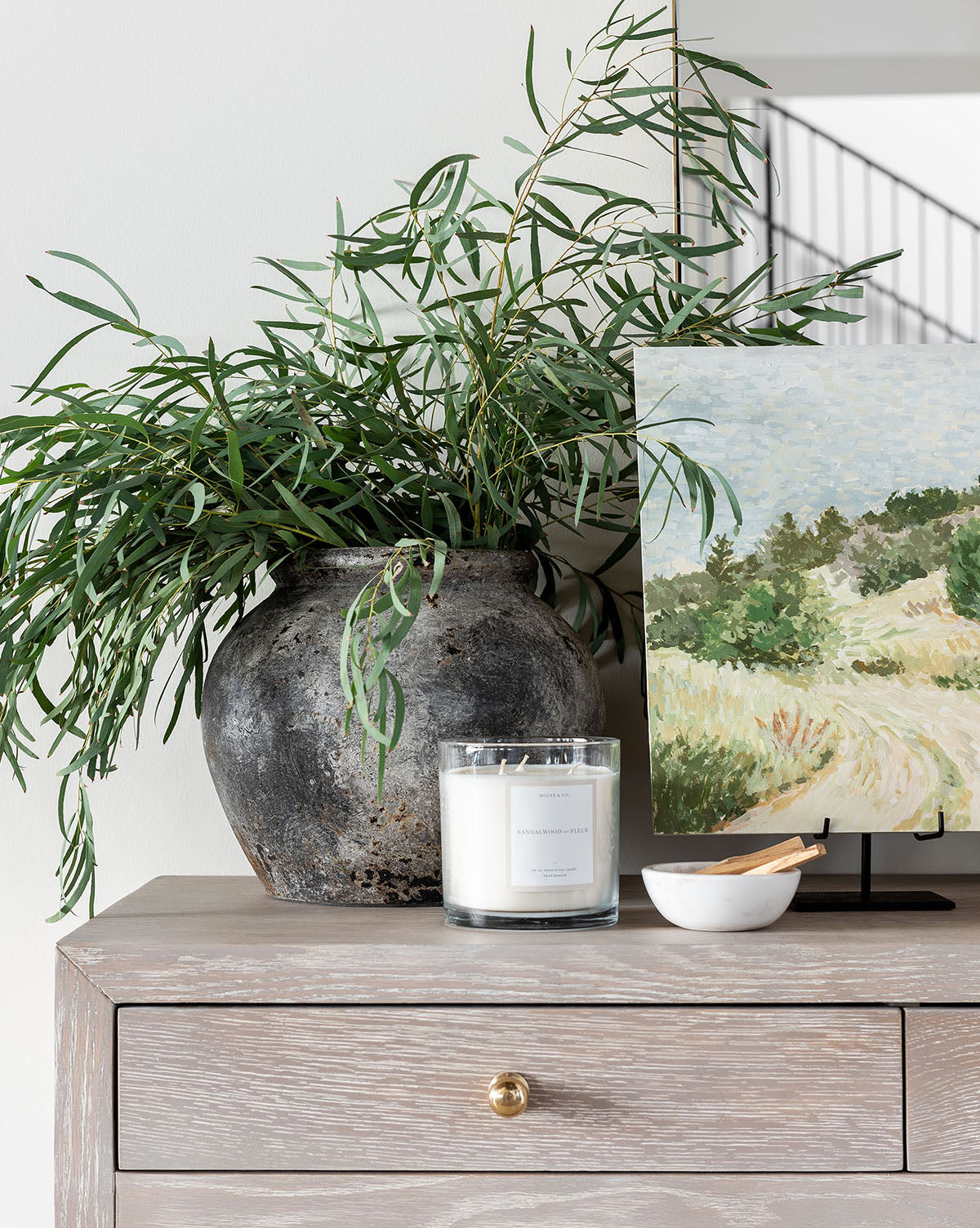 An Aged Terracotta Jar with green foliage, a white candle, and a small bowl of matches decorate a light wooden dresser beside a landscape painting, bringing textured home decor charm to the white wall.