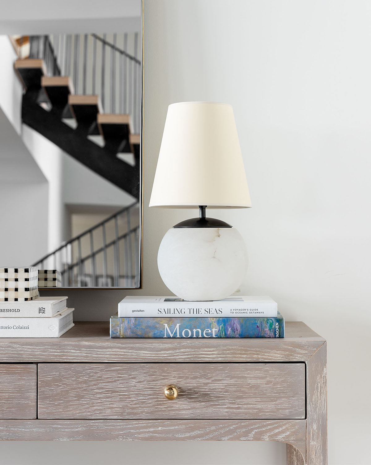 A Terri Round Table Lamp with a white shade and alabaster base sits on stacked books, including one titled Monet, on a wooden console table. Behind it, a large mirror reflects stairs with black railings, by McGee & Co.